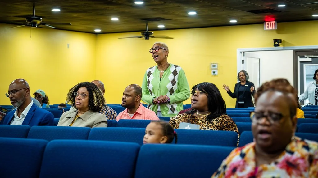 A woman stands and speaks in a room with yellow walls, while seated audience members listen. The room has blue chairs, ceiling fans, and an exit sign visible in the background.