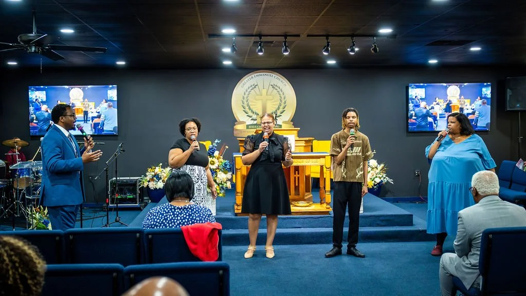 Five people standing on a stage leading a church service, with two screens displaying the same scene behind them. They are holding microphones, and there are flowers and musical instruments on the stage.