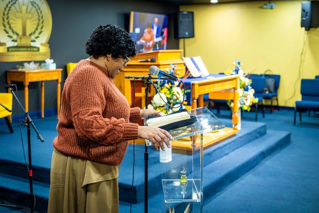 A woman in a brown sweater standing at a clear podium in a church or event hall, reading from a book or notes, with a microphone in front. Behind her are decorated tables with flowers, a religious emblem on the wall, and a television screen.