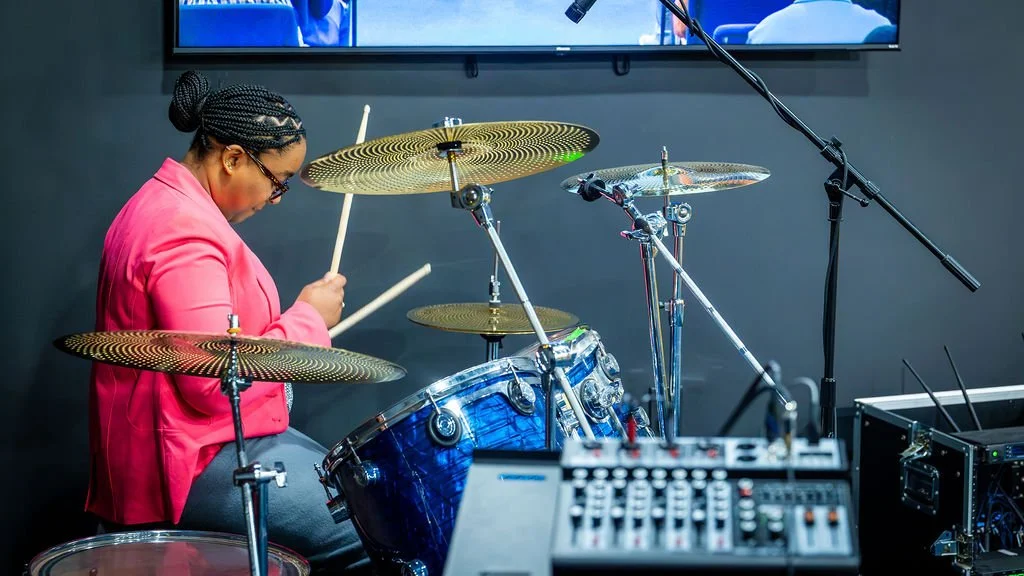 A woman playing a drum set, wearing a pink blazer and glasses, in a music studio with microphones and audio equipment.