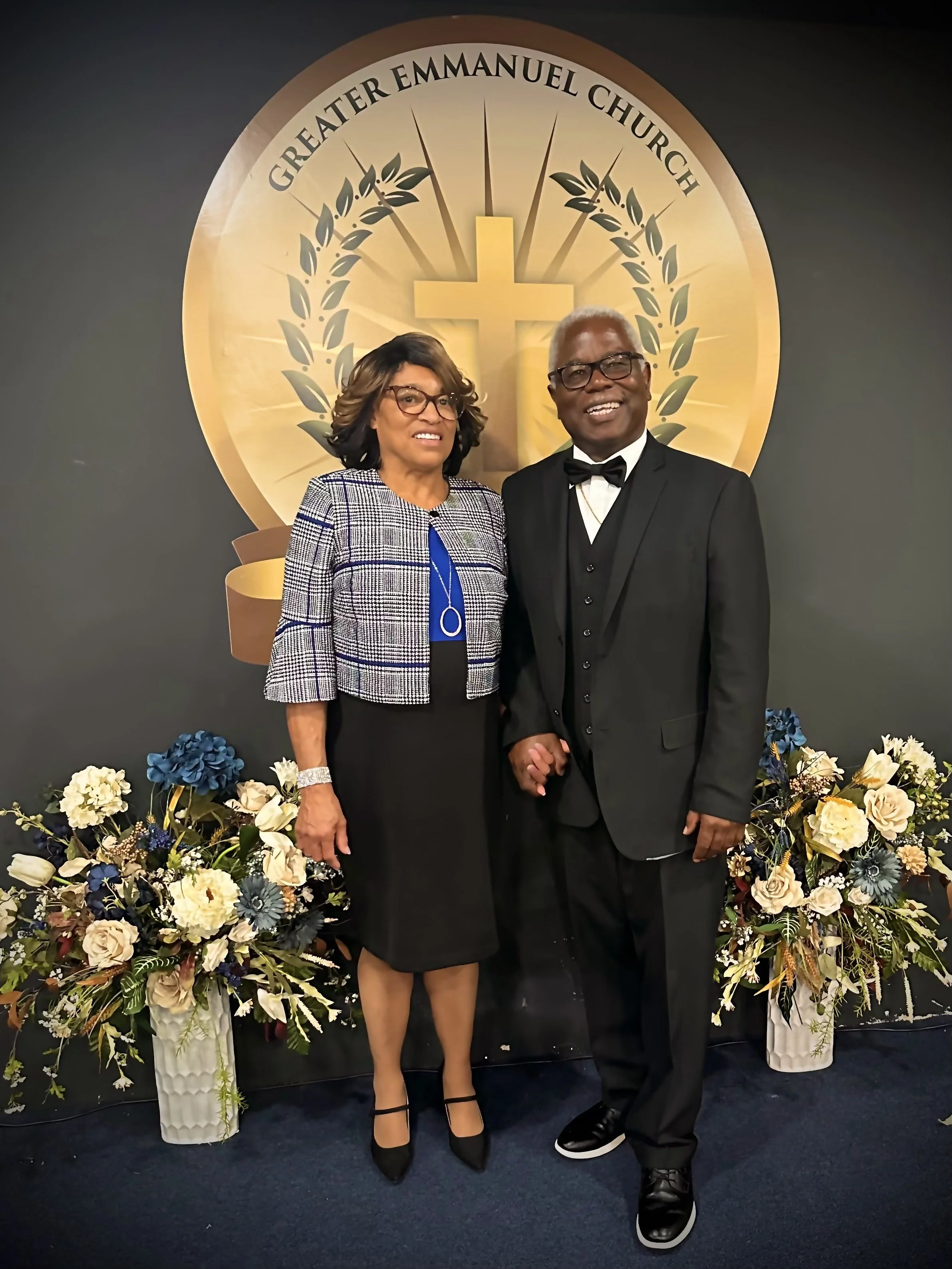 A man and woman dressed in formal attire standing in front of a backdrop with a cross and the words 'Greater Emmanuel Church'. They are holding hands and smiling, with floral arrangements on either side.
