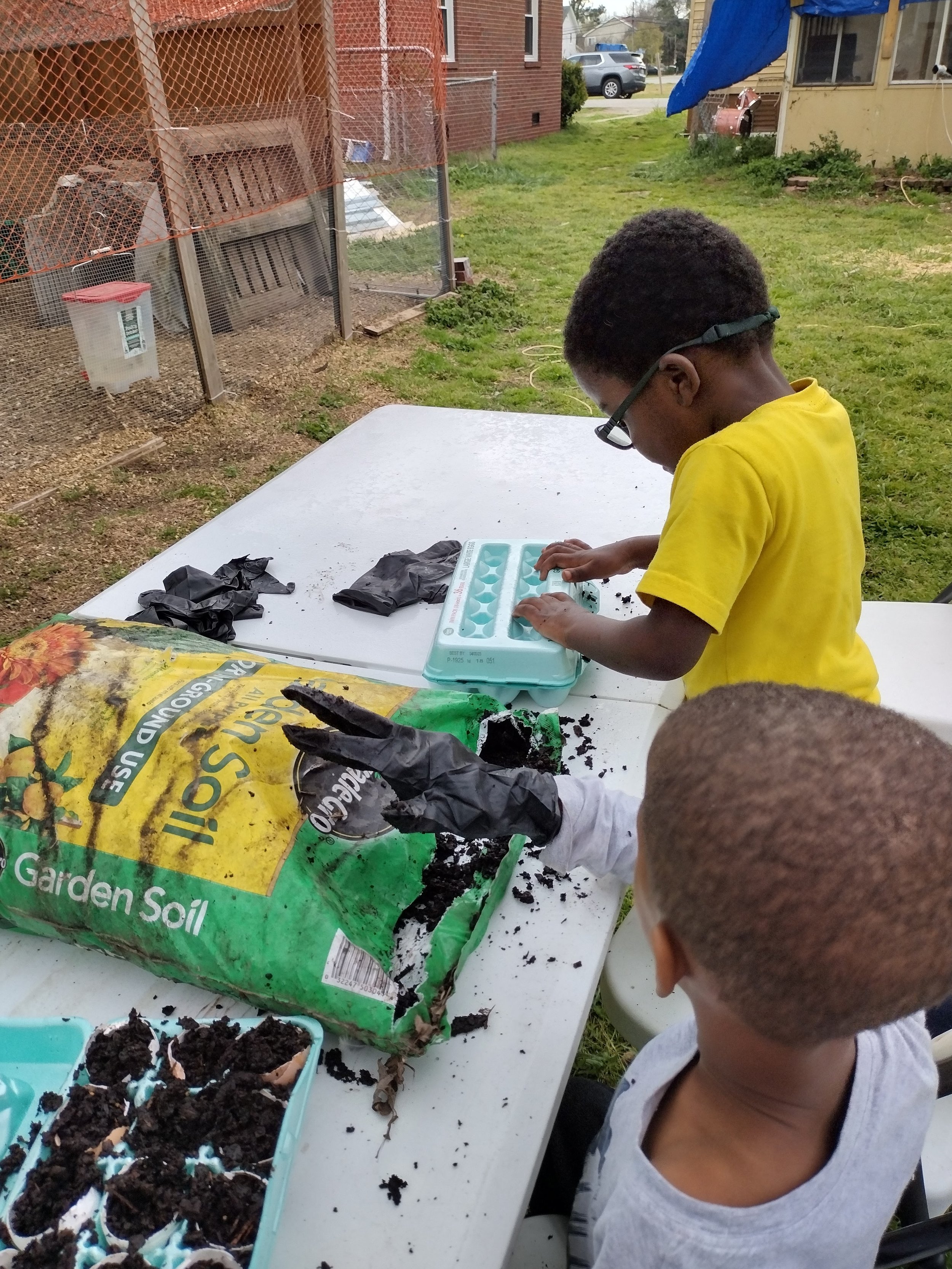 Two children planting seeds using an egg carton and gardening soil outdoors.