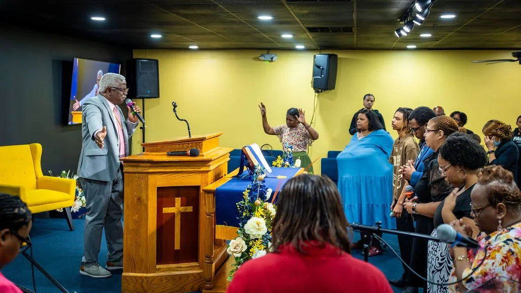 Man speaking at a church podium with a cross, with hands raised in prayer, surrounded by a congregation with eyes closed or hands raised, brightly lit with a yellow wall background.