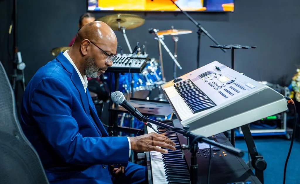 A man in a blue suit playing a keyboard in a music studio with a microphone in front of him, and drums and electronic equipment in the background.