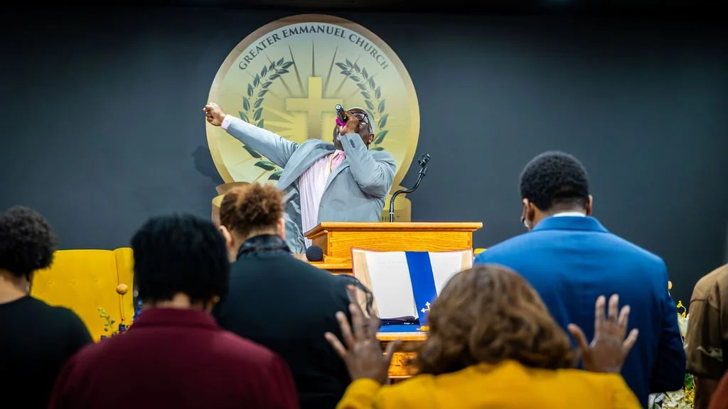 A person in a gray suit and pink shirt is speaking or singing into a microphone at a church podium. The church emblem with a cross and the words 'Greater Emmanuel Church' is visible on the wall behind them. Several people in the congregation are raising their hands, with some facing forward and others with their backs to the camera.