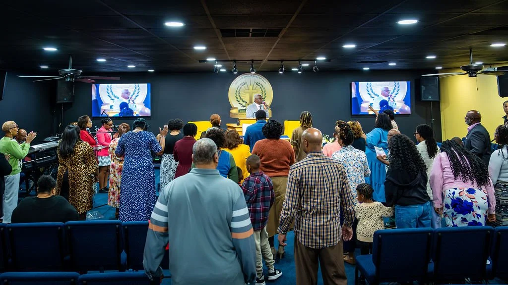 A church service with a congregation standing and clapping, led by a pastor at the front, with two screens displaying a speaker and a religious emblem.
