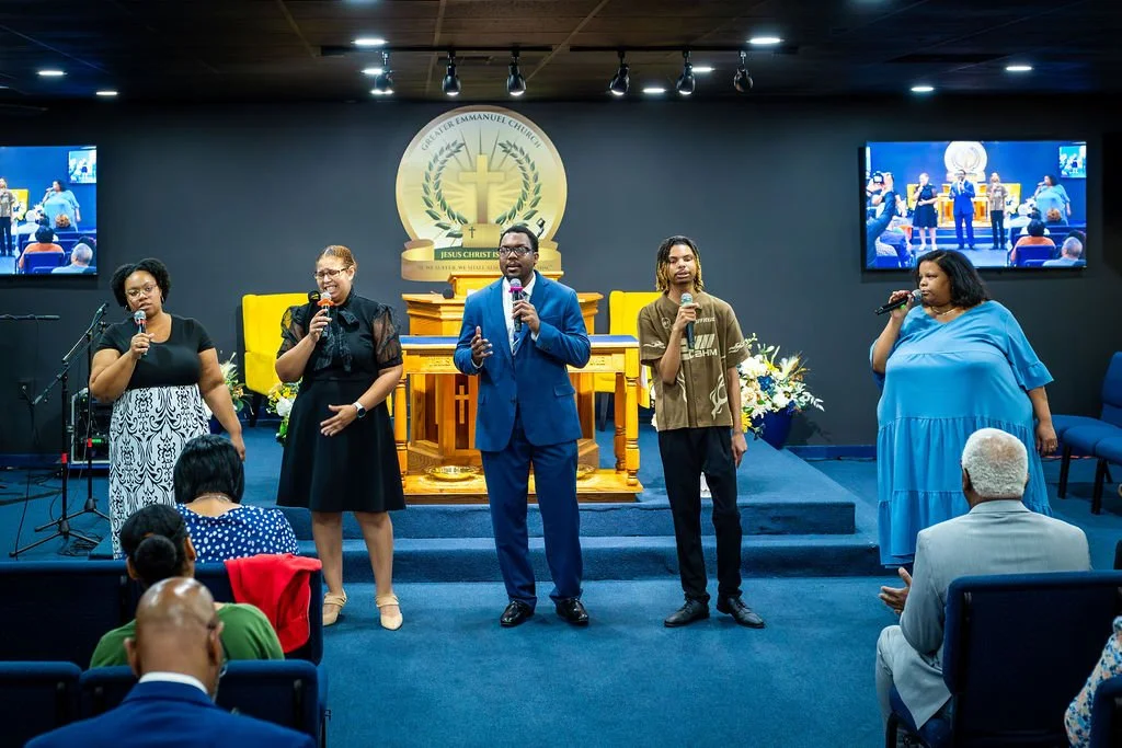 A diverse group of five people standing on stage in a church, singing into microphones during a service. The church's logo is visible behind them and there are large screens on either side showing their performance. Audience members are seated in front.