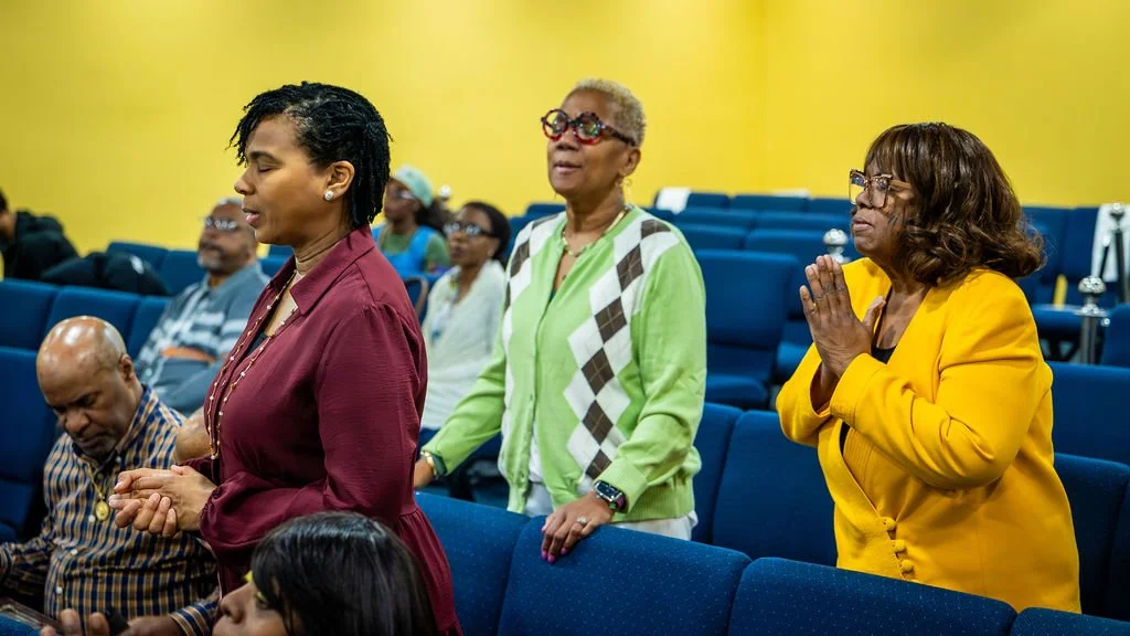 Group of people in prayer or meditation inside a church or meeting hall with blue seats and a yellow wall.