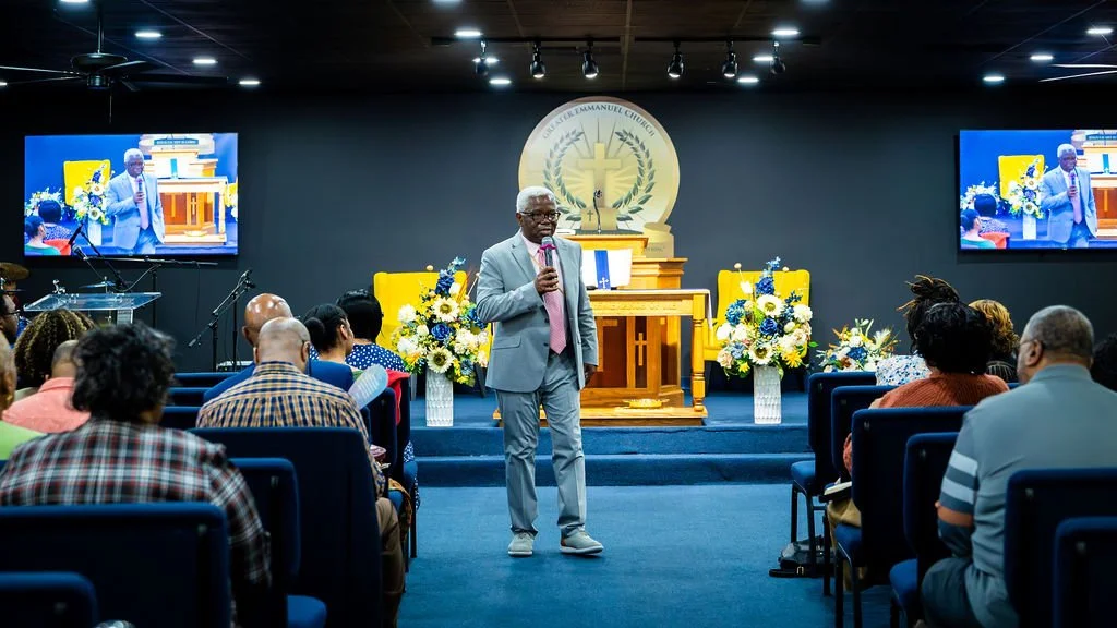 A church service with a man in a gray suit speaking into a microphone at the front of the church, flanked by floral arrangements, with an altar and a large church logo behind him. Two screens display the speaker and the congregation.