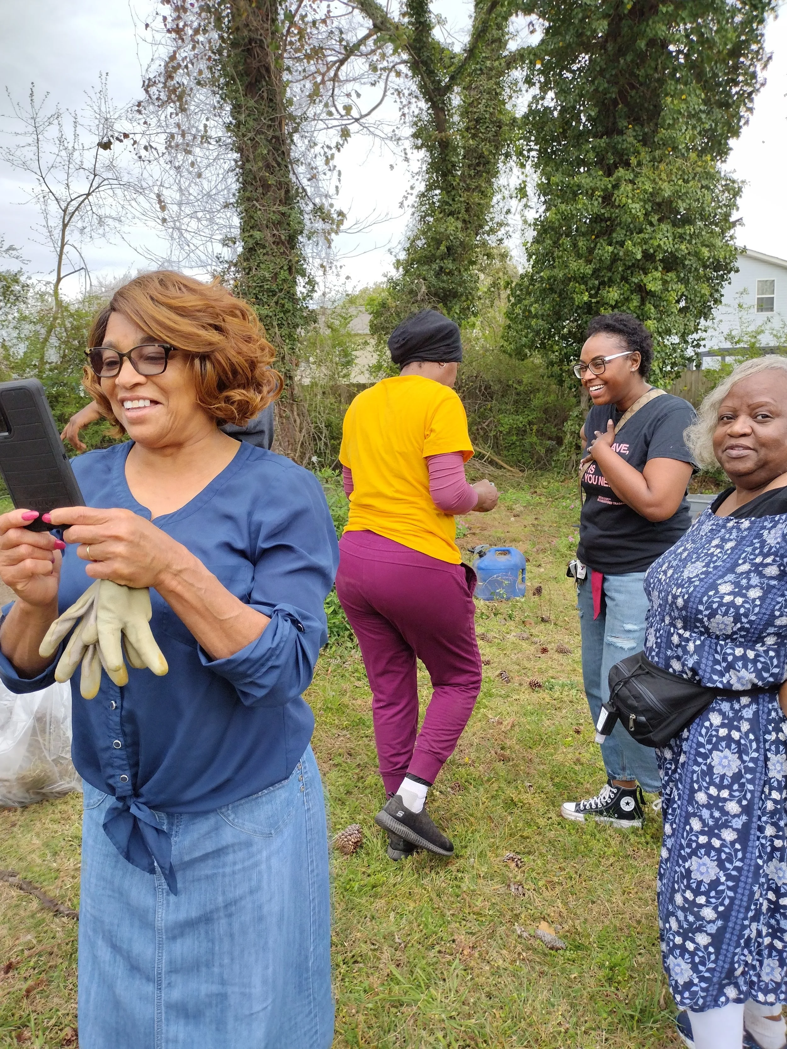Four women standing outdoors in a grassy area with trees in the background, one woman in front is looking at her phone, two women are talking and smiling, and one woman is standing with her hands folded, wearing glasses and a blue dress.