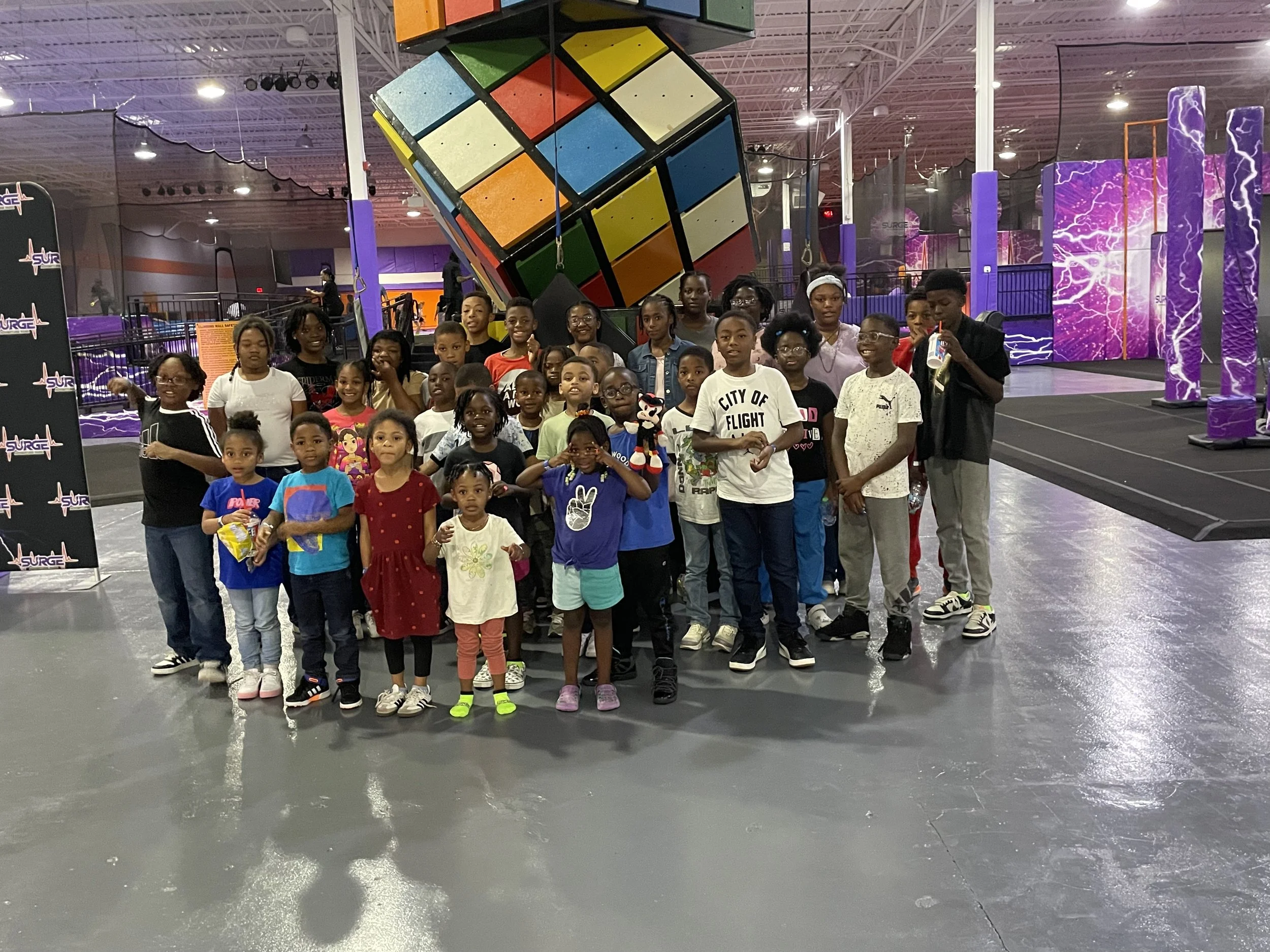 A group of children and a few adults posing for a photo inside an indoor amusement or trampoline park, with large colorful theming elements like a giant Rubik's Cube and purple lightning-themed structures in the background.
