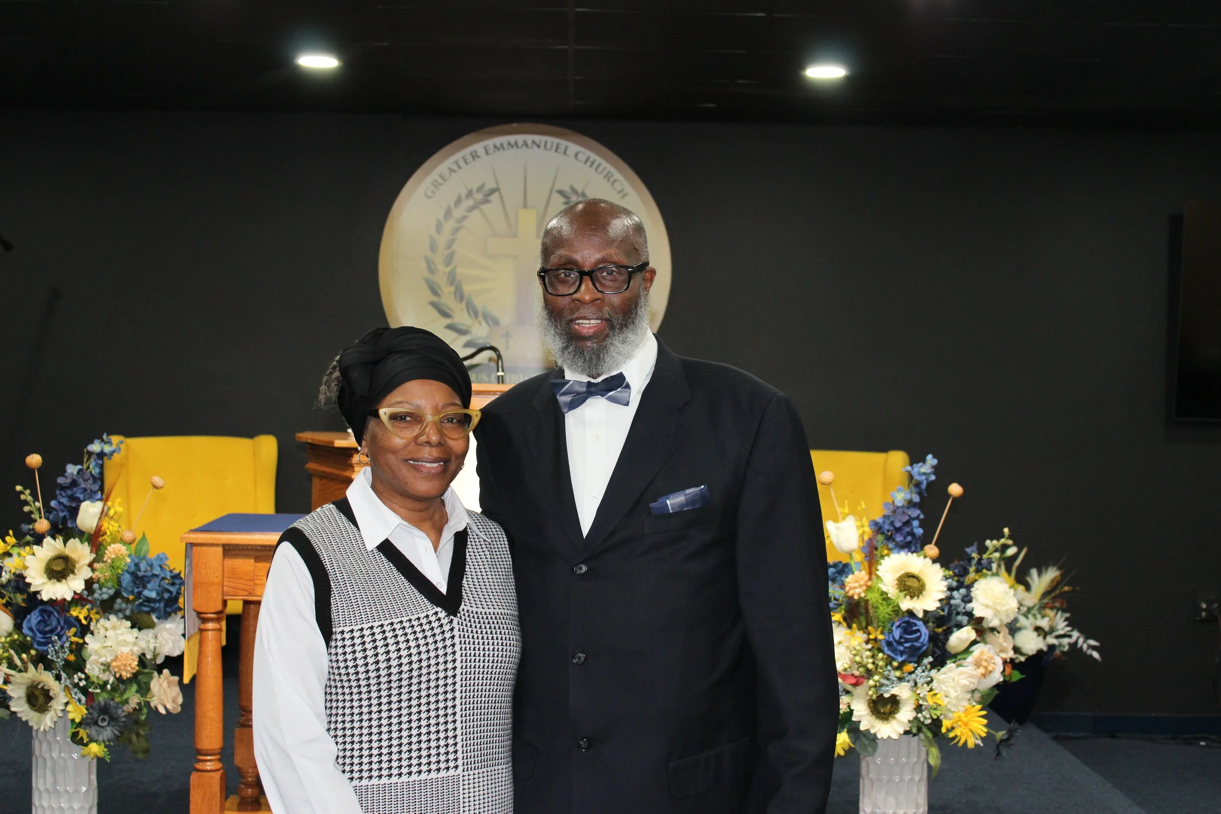 A man and woman posing together in a church setting with floral arrangements, a podium, and a church emblem in the background.