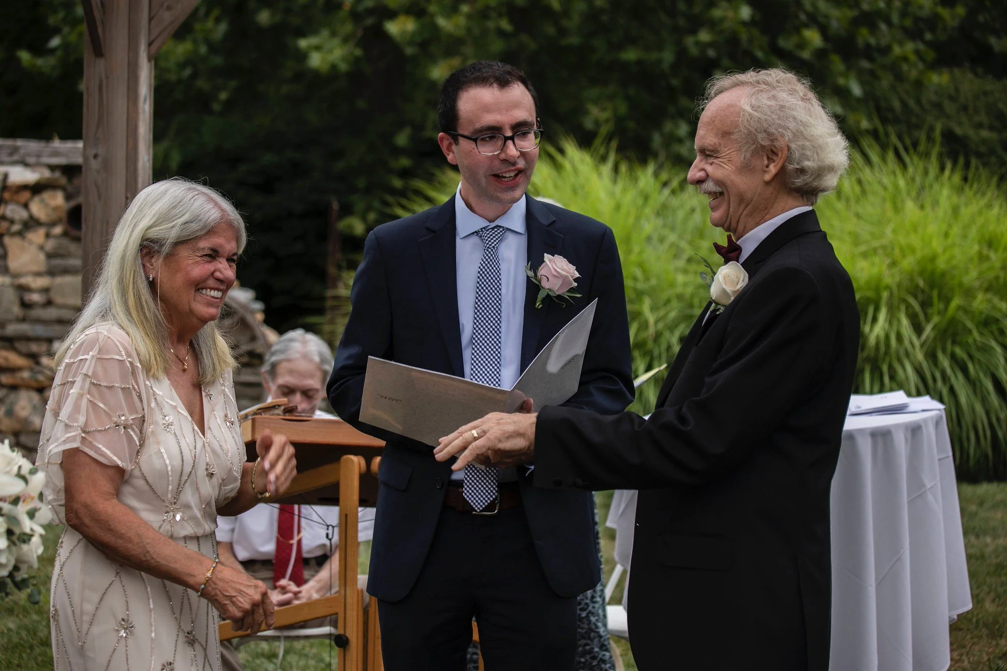 A wedding ceremony outdoors with four people, including a bride and groom, during the ring exchange. The groom is in a dark suit, the bride is not fully visible. An officiant and an older woman are smiling in the background, with lush green plants and a stone structure behind them.