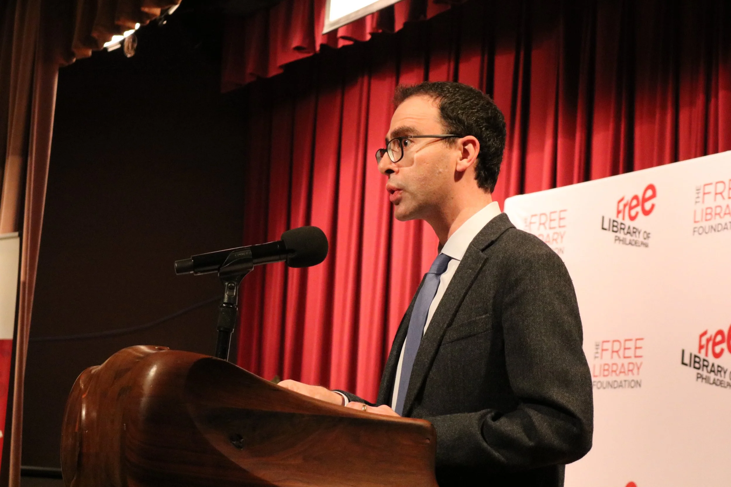 Man in a suit and glasses speaking at a podium in front of a red curtain and a backdrop with 'Free Library of Philadelphia' logos.