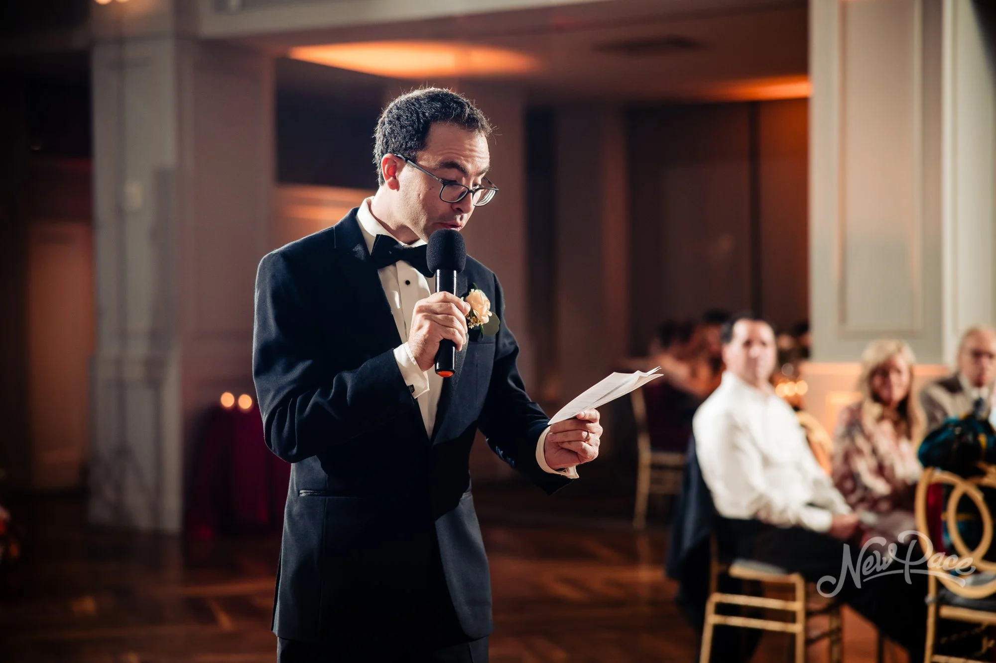 A man in a tuxedo giving a speech at a wedding reception, holding a microphone and reading from a paper, with guests seated at tables in the background.