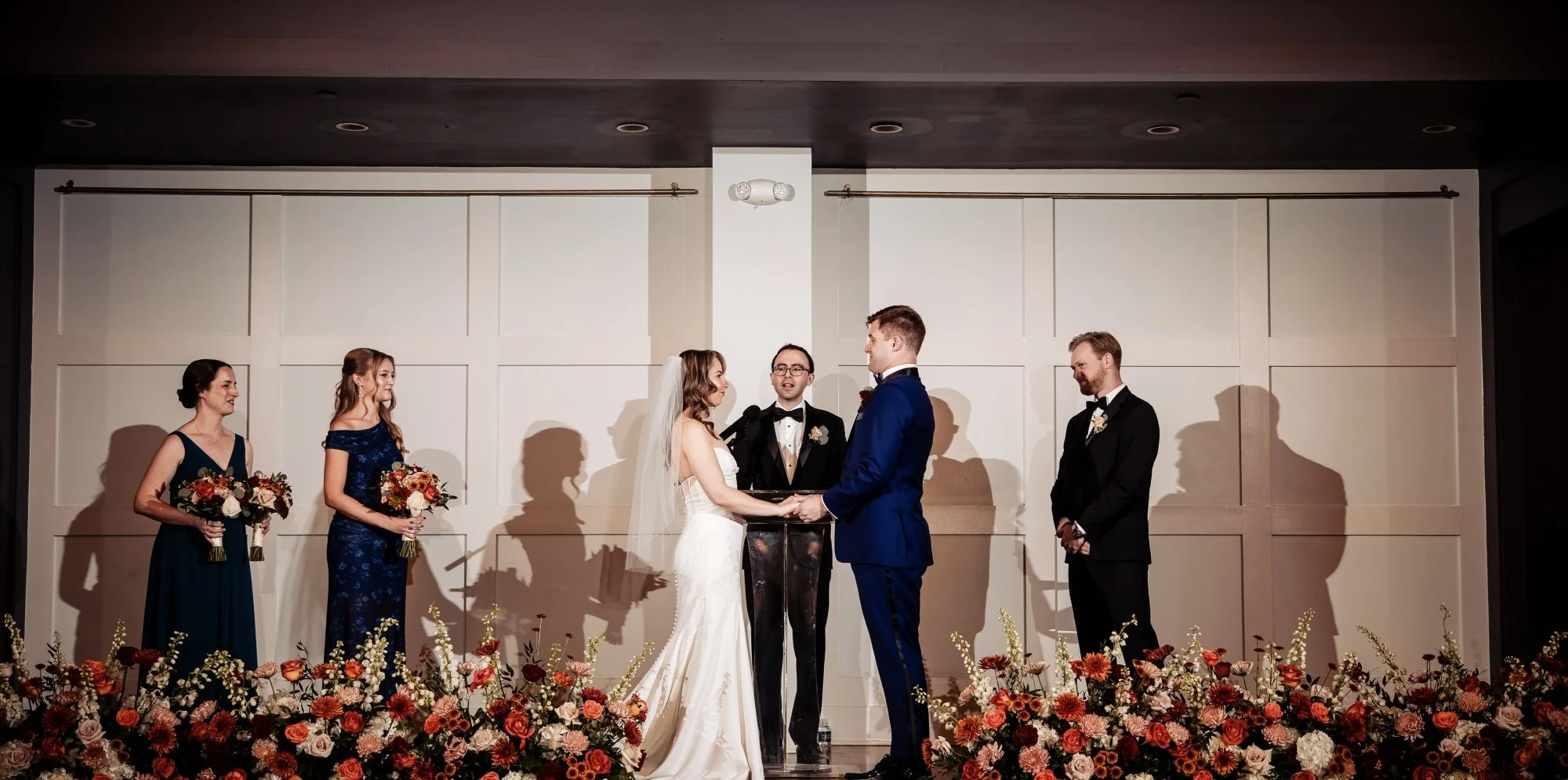 A bride and groom exchange vows at their wedding ceremony, surrounded by bridesmaids holding bouquets, officiant, and groomsman, with floral arrangements in the foreground.