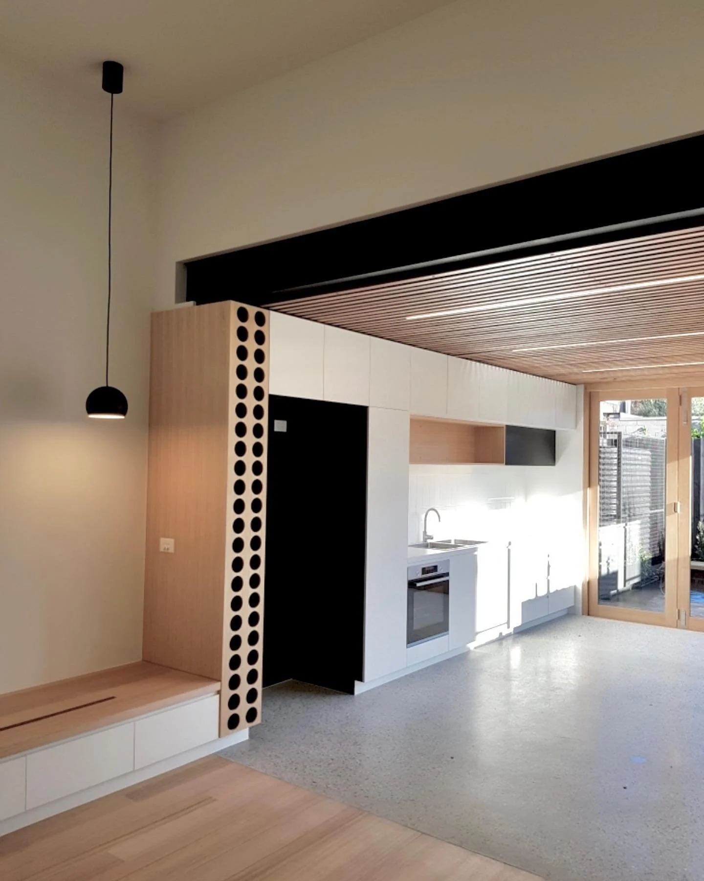 Modern kitchen with white cabinets, built-in oven, and wooden ceiling, near large glass door opening to outdoor space.