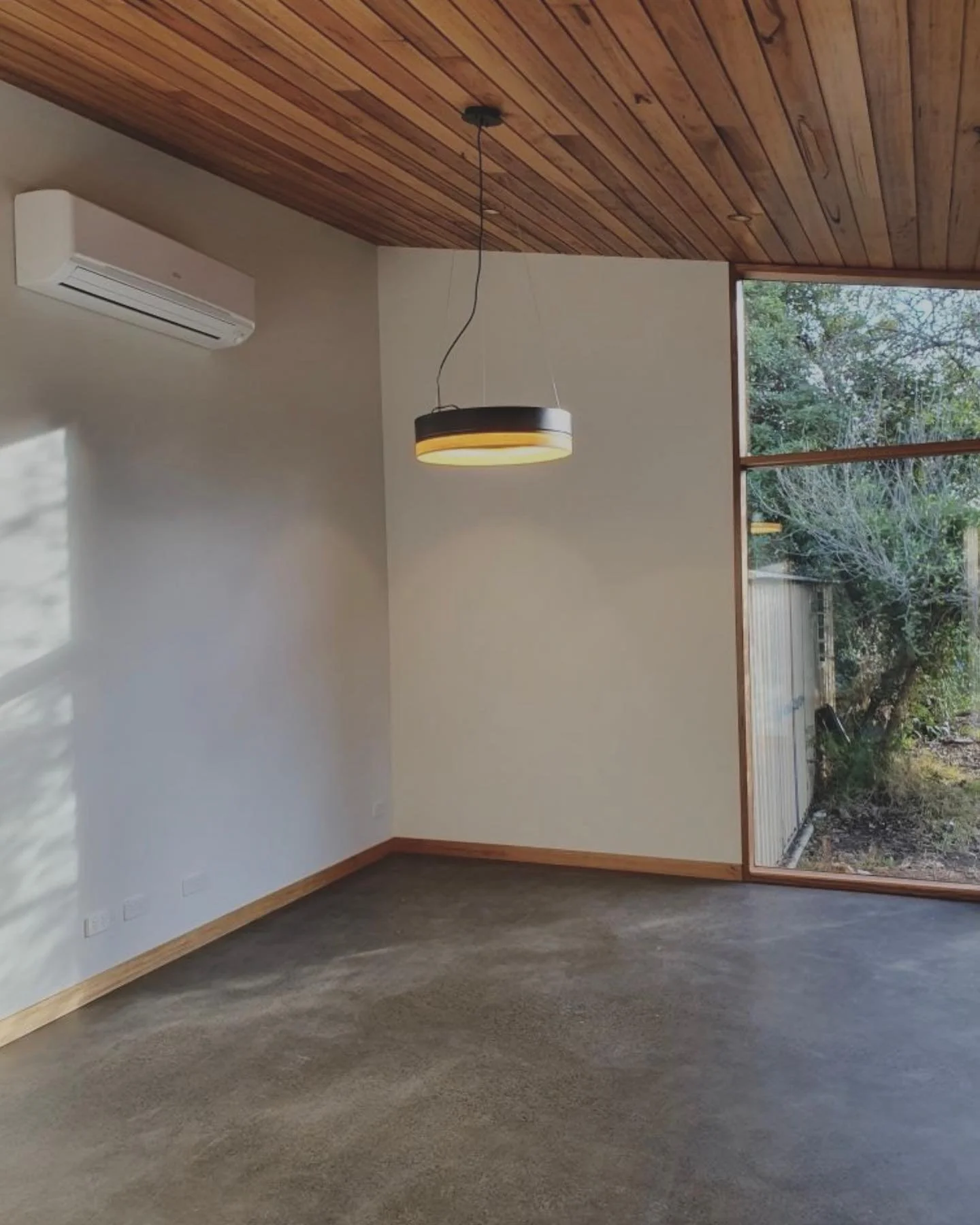 Empty room with wood-paneled ceiling, large window, wall-mounted air conditioning unit, hanging light fixture, and concrete floor.