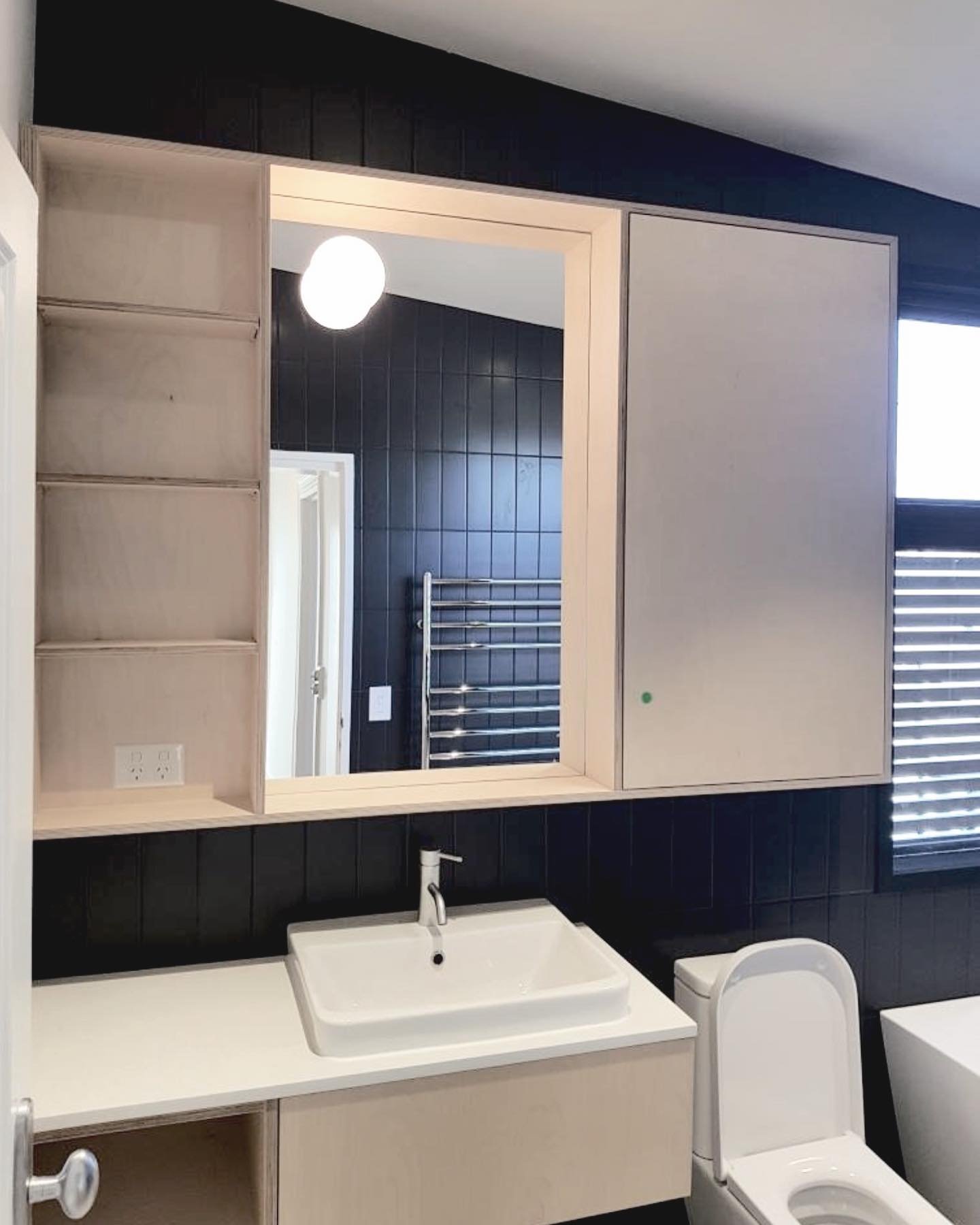 Modern bathroom with light wood vanity, white rectangular vessel sink, black tiled wall, and large mirror, with a toilet to the side and window with blinds.