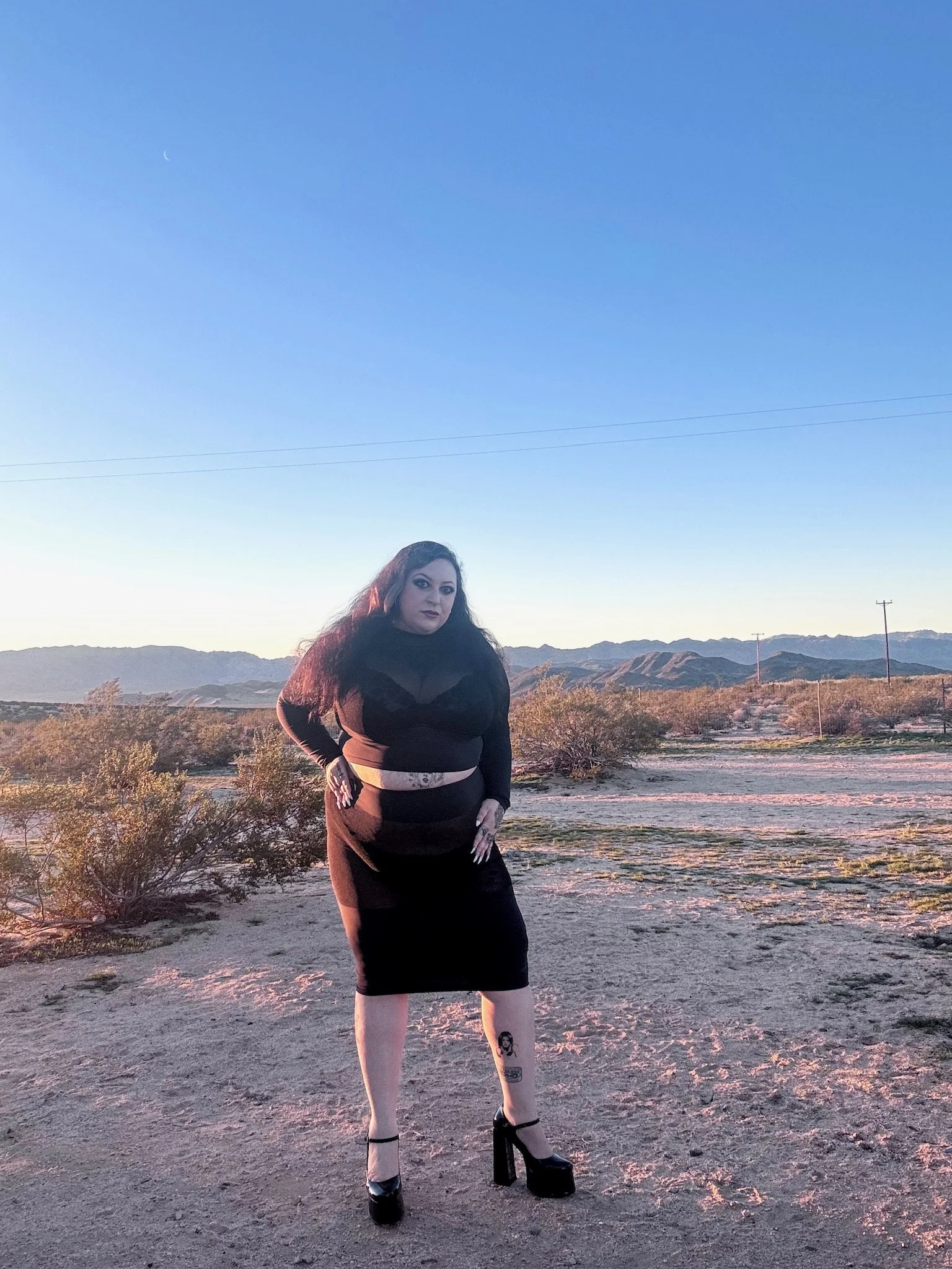A woman with dark hair and tattoos wearing a black dress and high heels standing outdoors in a desert landscape during sunset with mountains in the background.