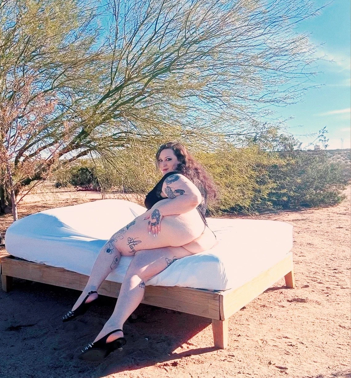 A woman with tattoos sitting on a mattress outdoors in a desert landscape with sparse trees and a clear blue sky.