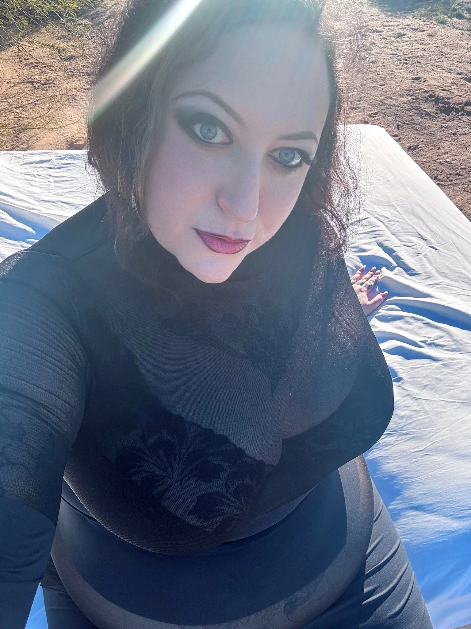 A woman with black hair and blue eyes taking a selfie outdoors on a sunny day, with a rocky ground background, wearing a black top with sheer lace details.