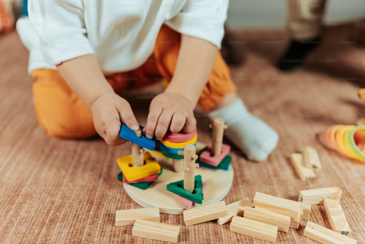 A toddler wearing an oversized white shirt, orange pants, and white socks is playing with colorful geometric wooden stacking blocks on a textured brown carpet.
