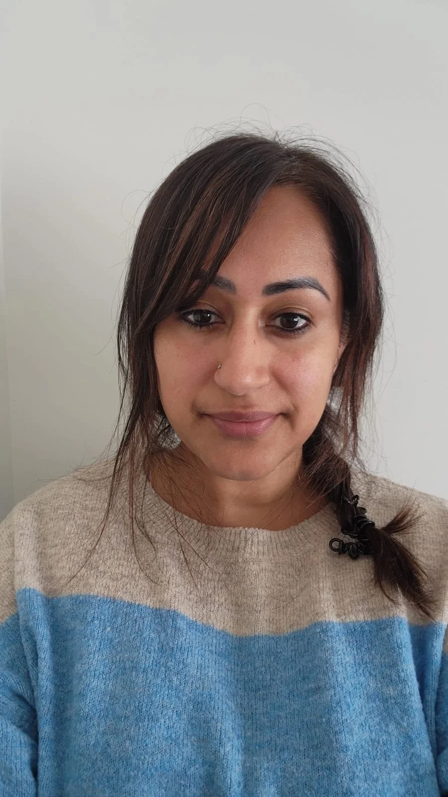 A woman with brown hair styled in a loose braid, wearing a beige and blue striped sweater, standing against a plain light-colored wall.