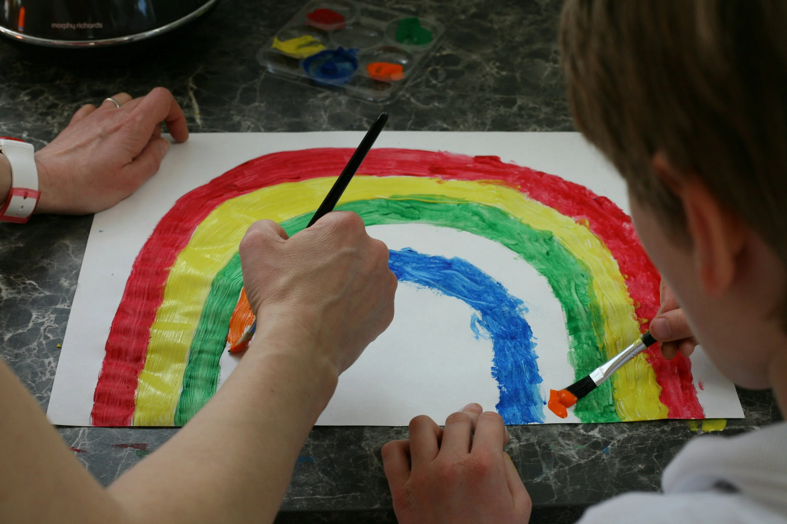 Two people painting a rainbow with brushes on a white sheet of paper, with a palette of paint nearby on a dark marble surface.