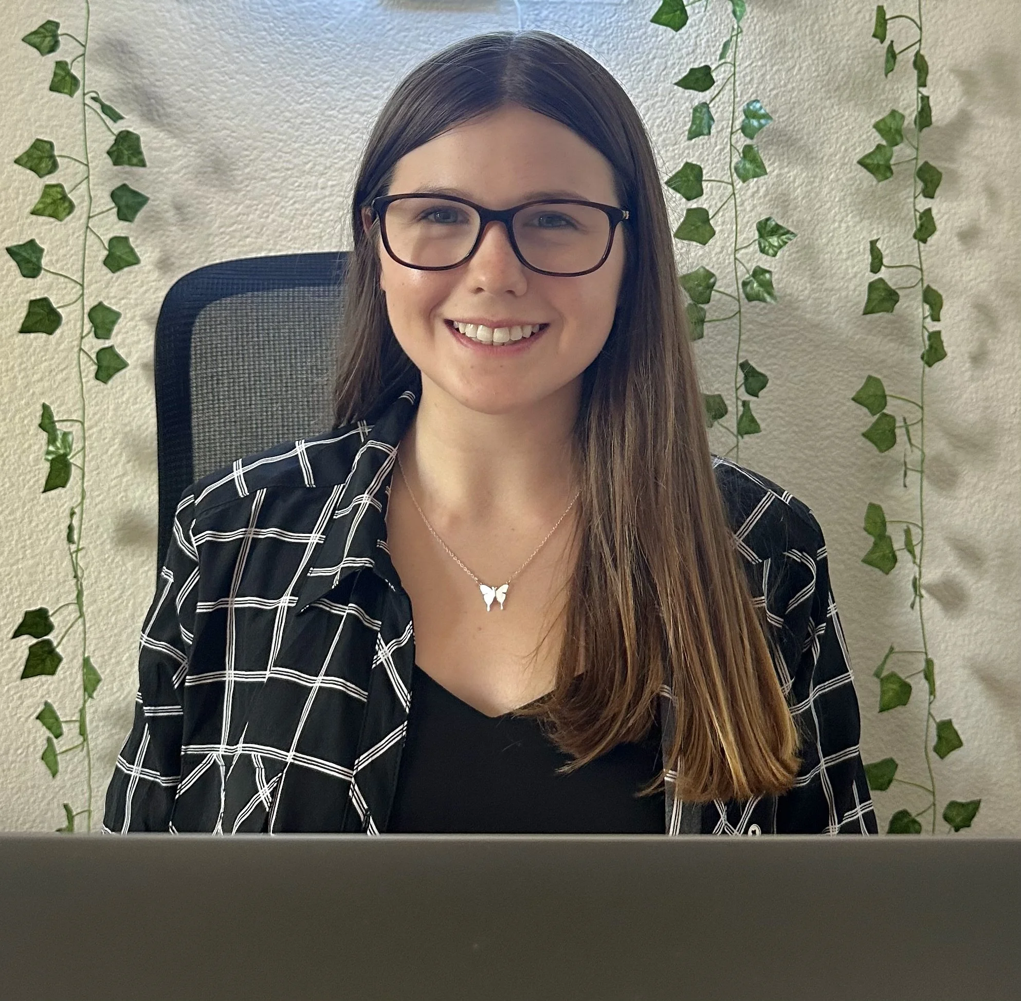A young woman with long brown hair, wearing glasses and a butterfly necklace, smiling at the camera while seated in an office chair with a computer in front of her. The background has a wall decorated with green ivy vines.
