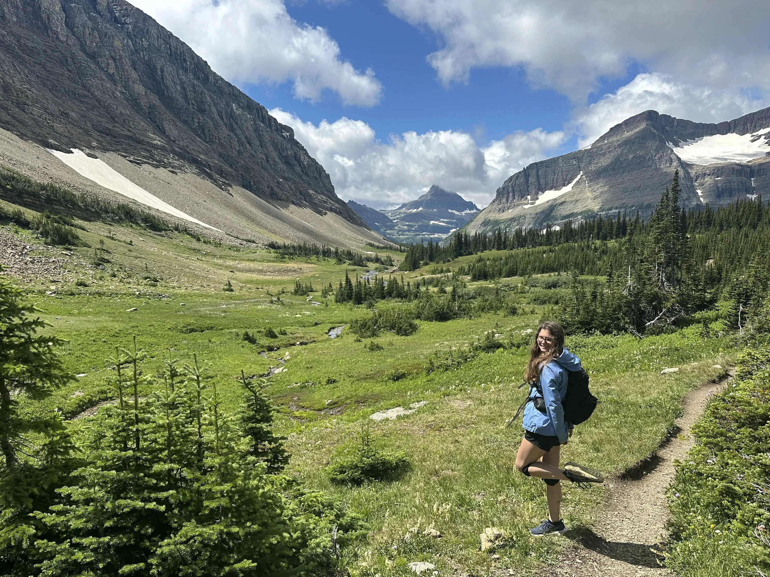 A woman in outdoor gear standing on a trail in a green valley with mountains, some snow, and a partly cloudy sky in the background