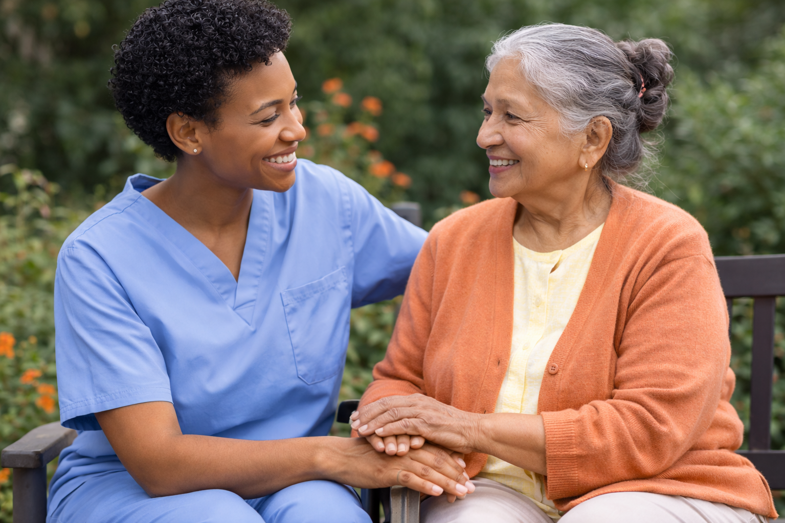 A young nurse in blue scrubs smiling and holding hands with an elderly woman with gray hair in a bun, wearing an orange cardigan, sitting on a bench outdoors surrounded by green foliage and flowers.