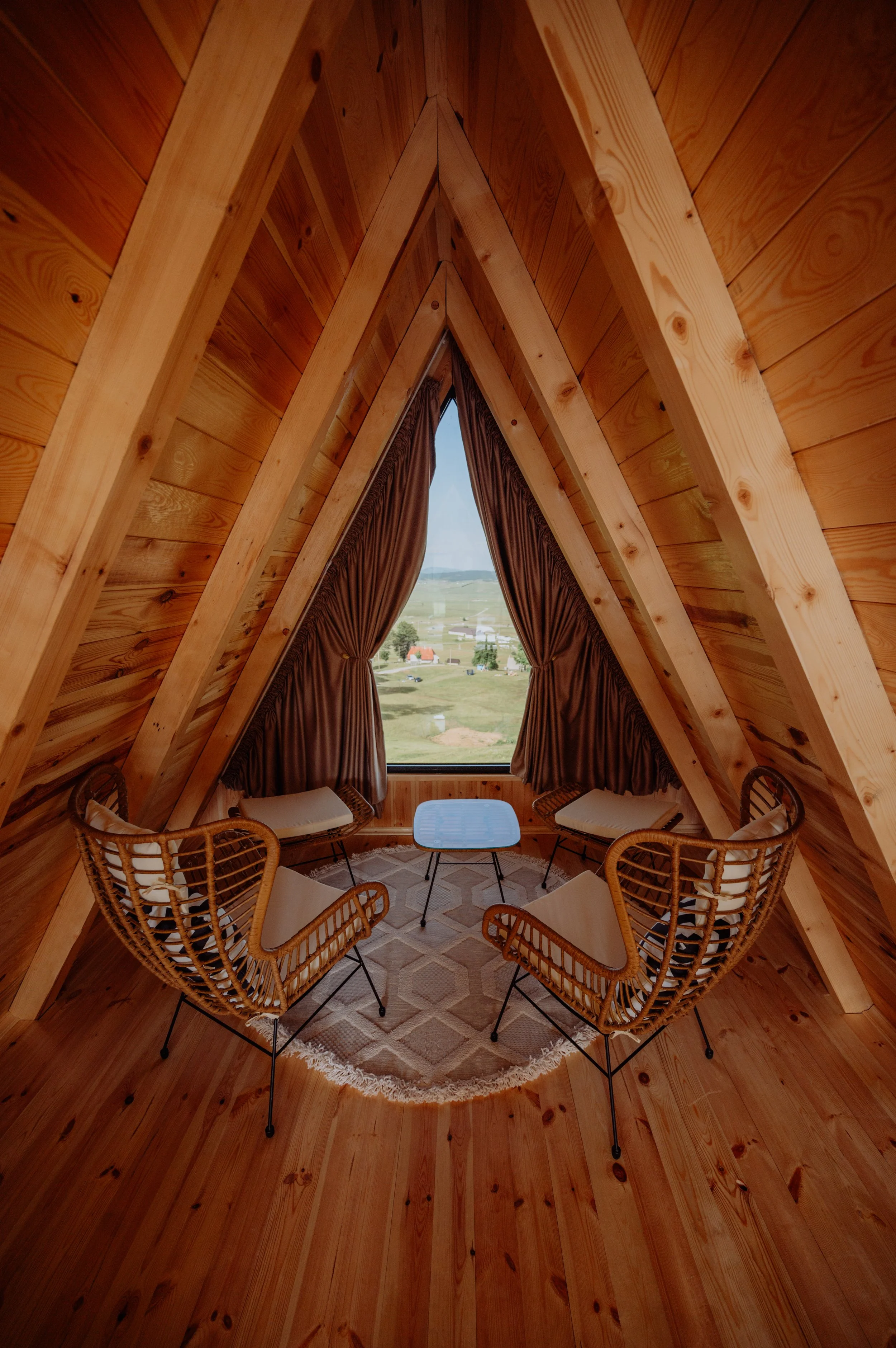 A cozy teepee with wooden interior, four rattan chairs with cushions, a small table in the center, a patterned rug, and a large window with brown curtains showing a rural landscape outside.