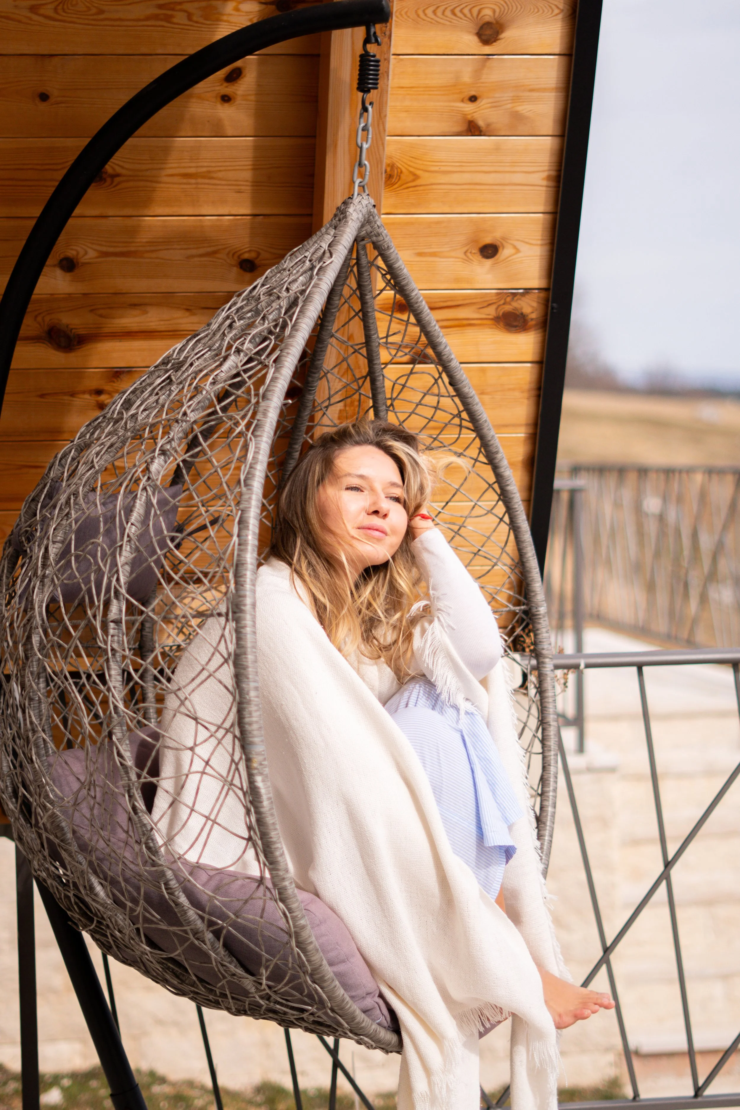 A woman sitting on a hanging wicker chair outdoors, with a wooden wall and railing in the background.