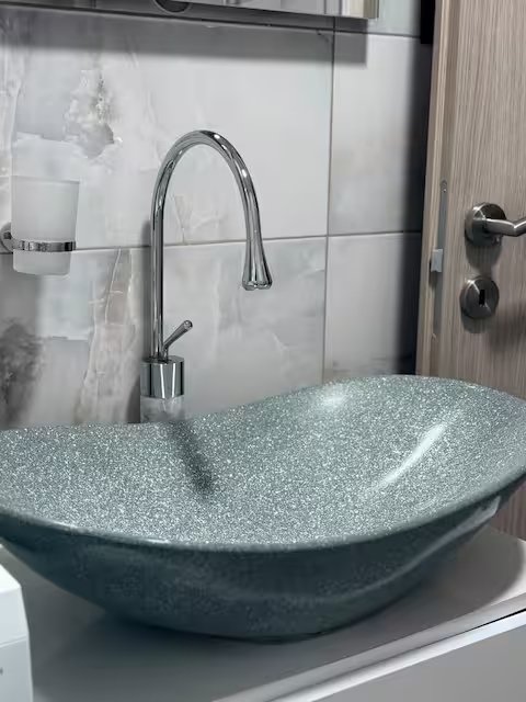 A modern bathroom sink with a textured, glossy finish and a curved, elongated shape sits on a white counter. There is a chrome faucet with a tall, curved spout above the sink. The background features white tiled walls and a wooden door with a silver 