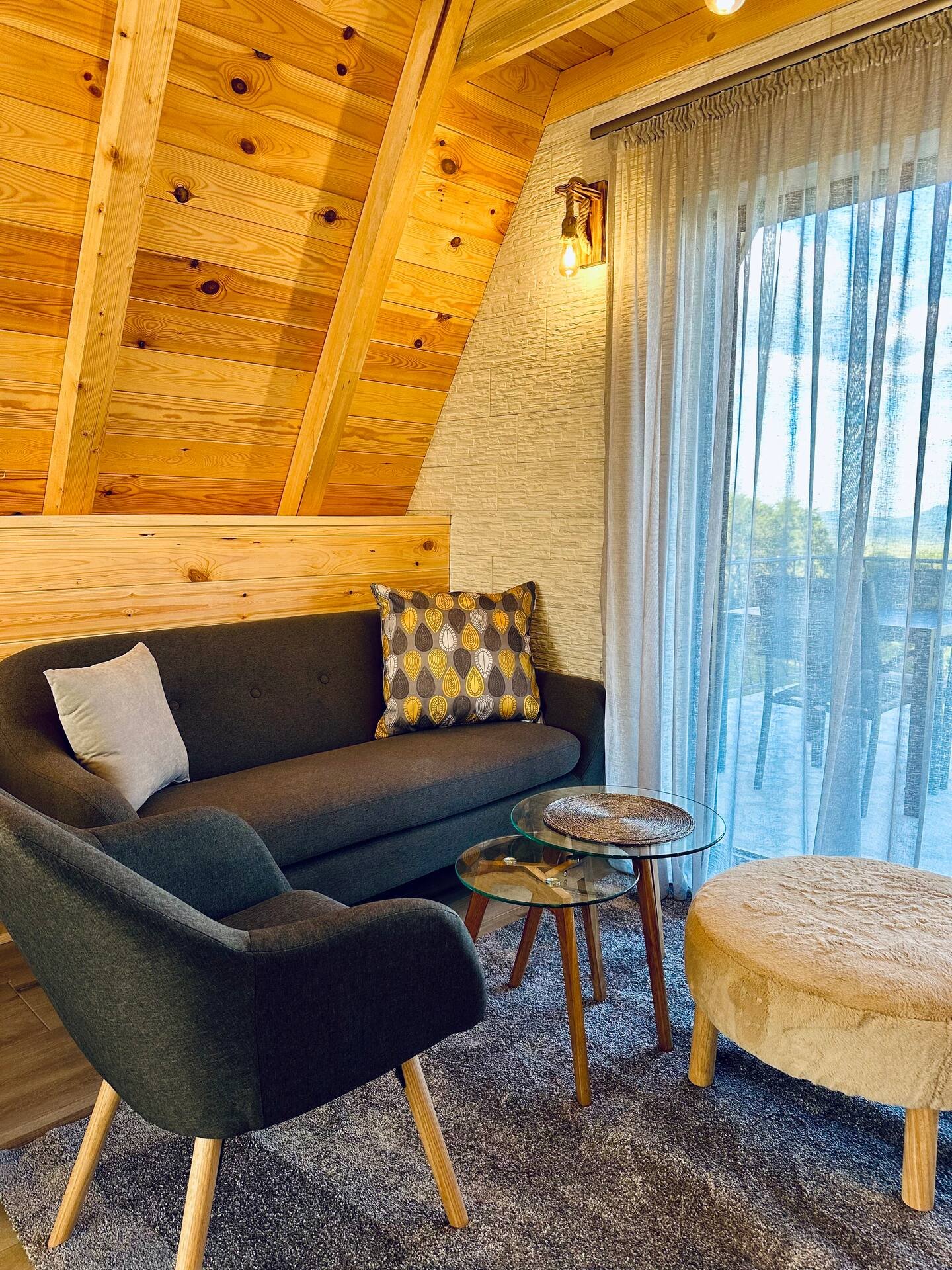 A cozy living room with a black sofa, a matching armchair, glass-top tables, a beige round ottoman, and a large window with sheer curtains. The room has wooden walls and ceiling, with warm lighting.