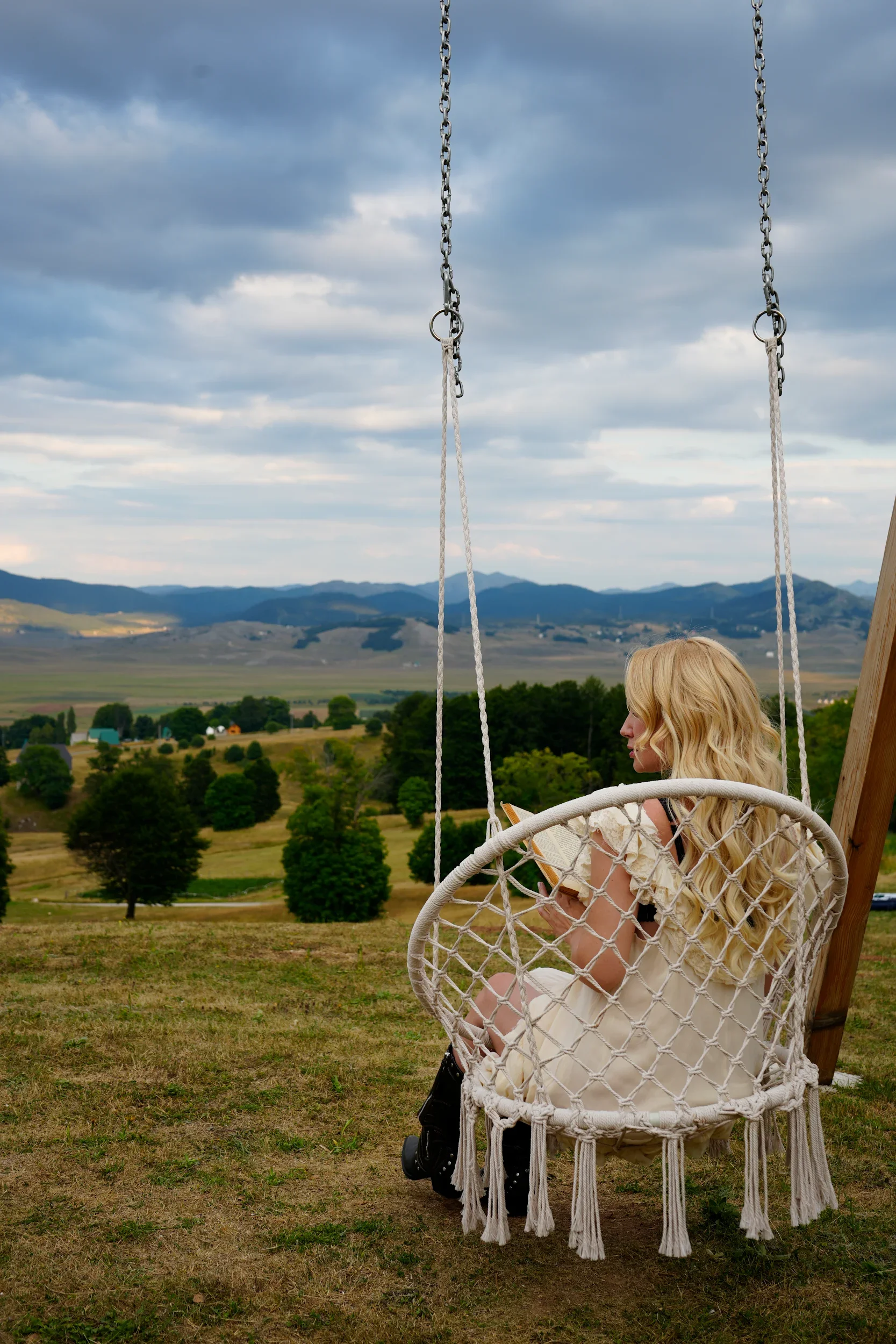 A woman with long blonde hair sitting on a hanging woven round swing on a grassy hill, reading a book. The background features a scenic view of rolling hills, trees, and a cloudy sky.