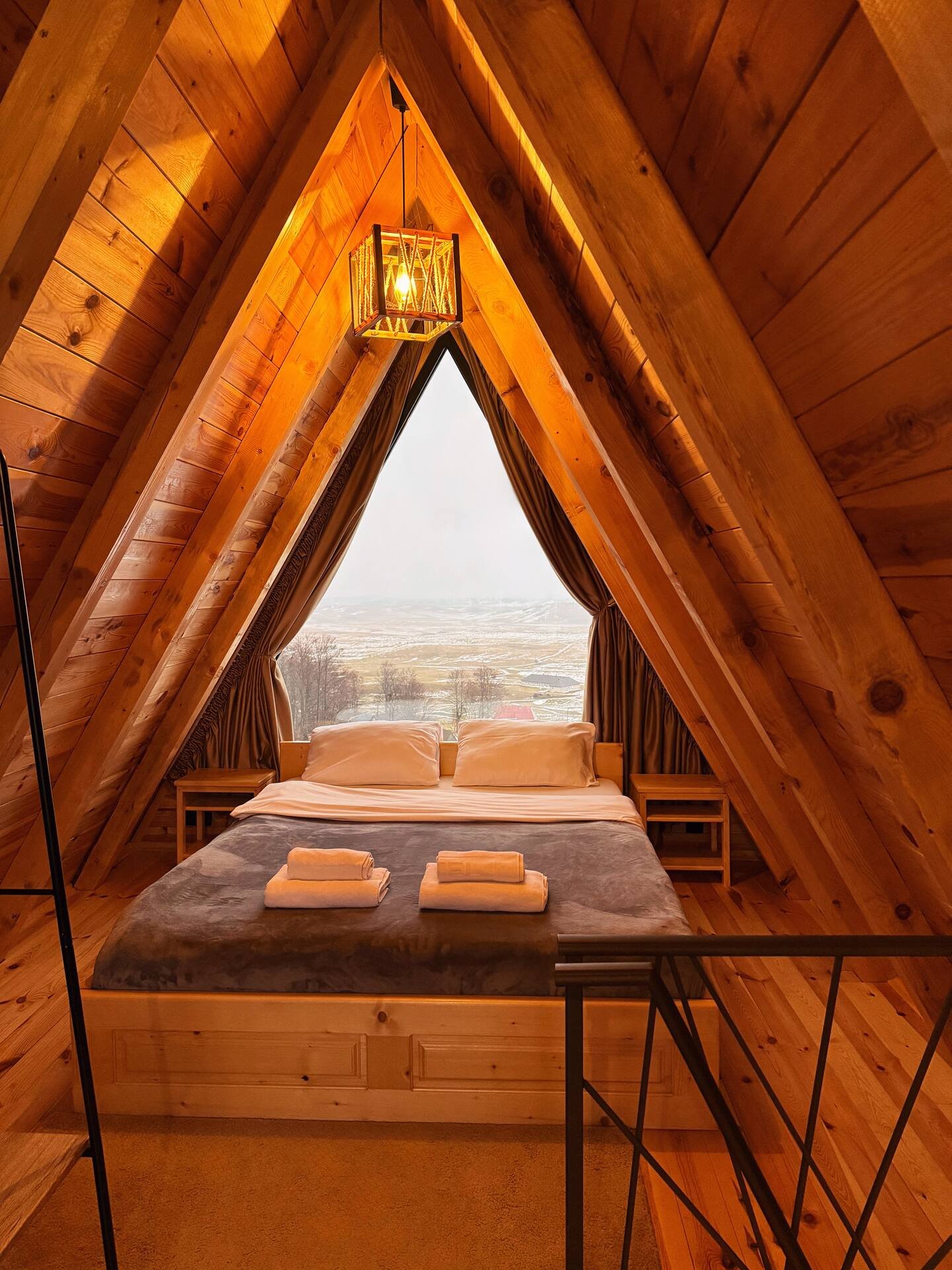 A cozy triangular A-frame wooden bedroom with a large bed, two pillows, folded towels, nightstands, and a large window showing a snowy landscape outside.
