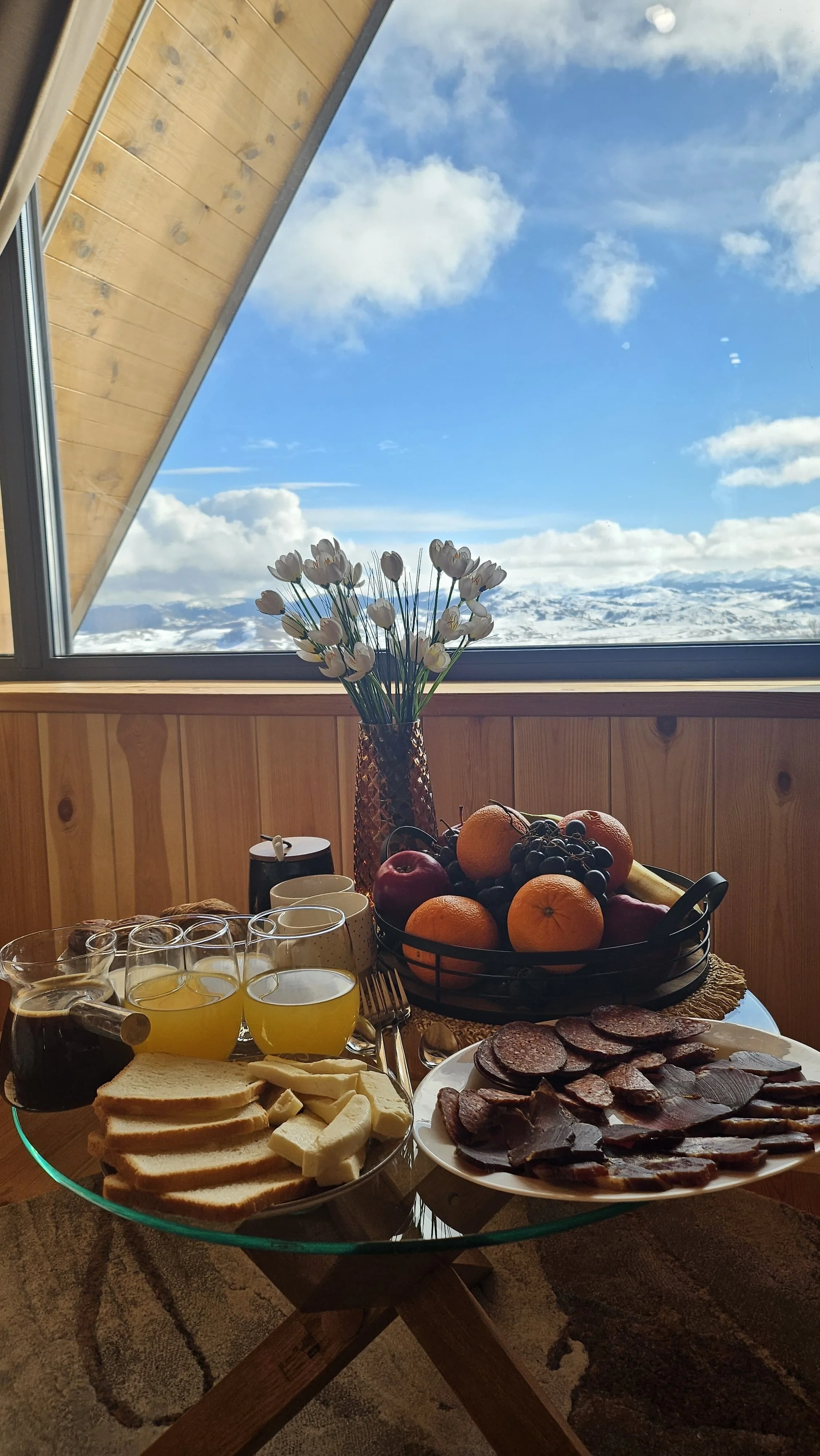 A table set with bread, cheeses, juice, and meats, with a bowl of fruit and a vase of white flowers, overlooking snowy mountains through a large window.