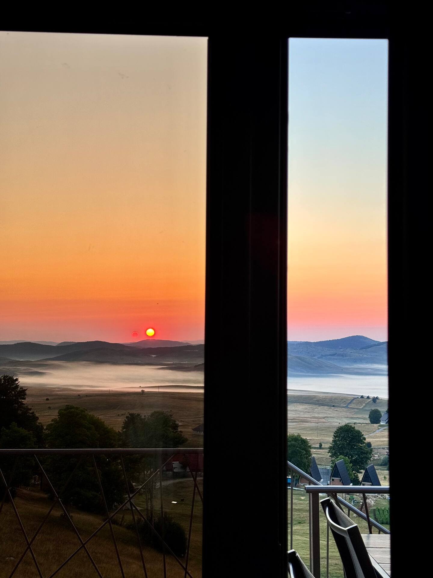 Sunset view through a window with two panes, showing a landscape with mountains, trees, and fog over a field, with the sun near the horizon.