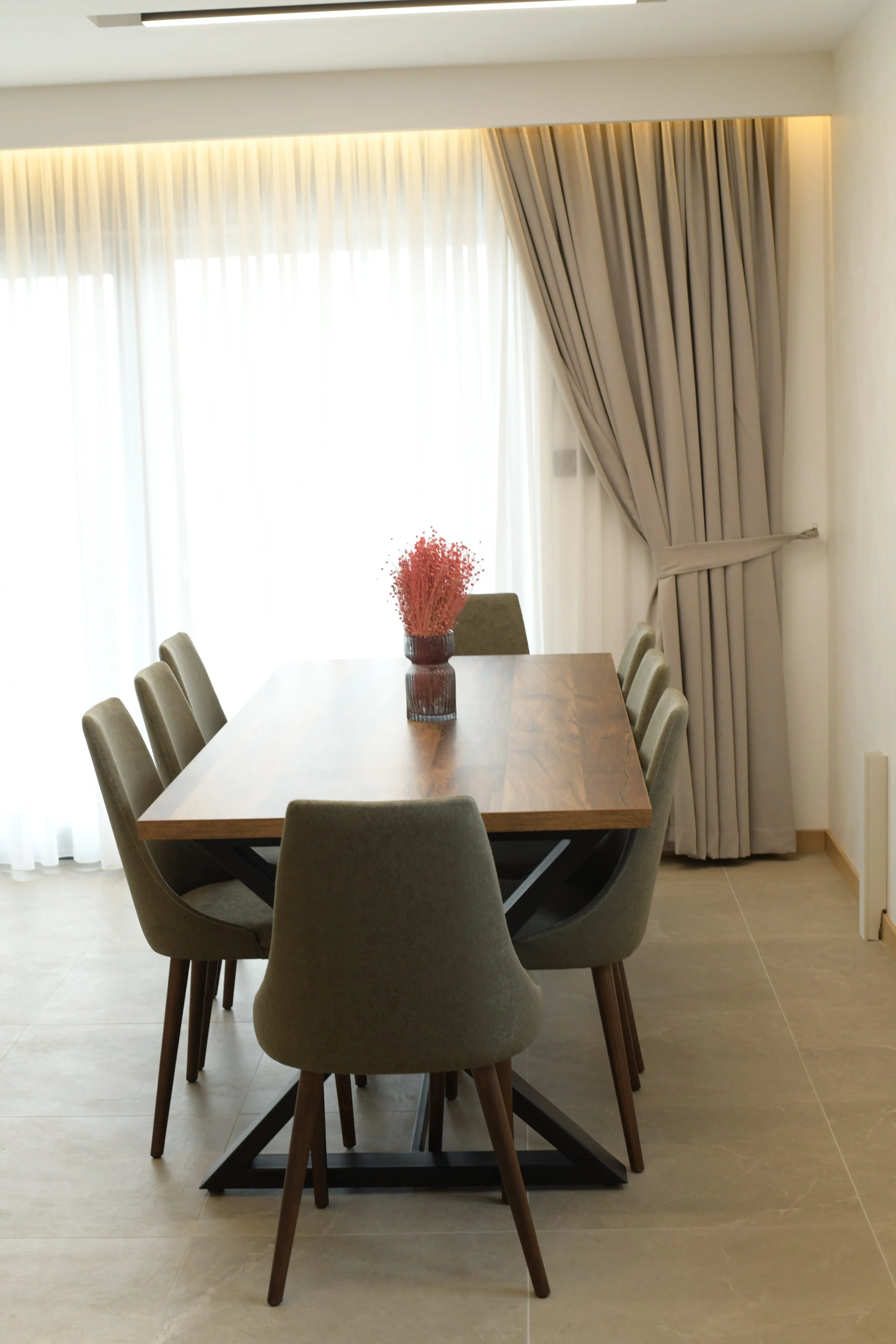 Dining room with six green upholstered chairs around a wooden table, a red flower arrangement in a glass vase, and beige curtains with sheer white curtains behind them.