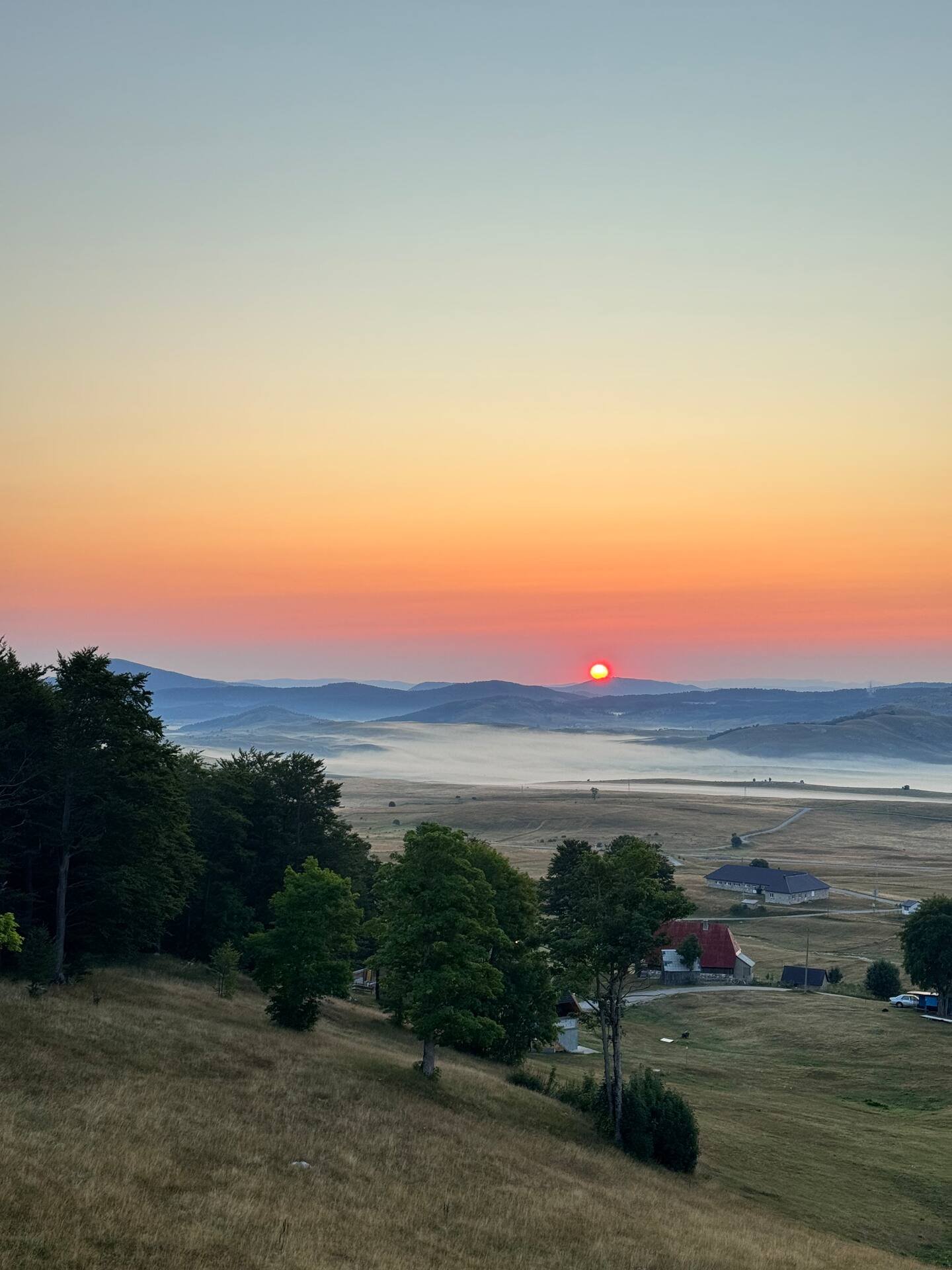 Sunset over rolling hills with scattered trees and a rural landscape, mist in the distance, and a few buildings visible.
