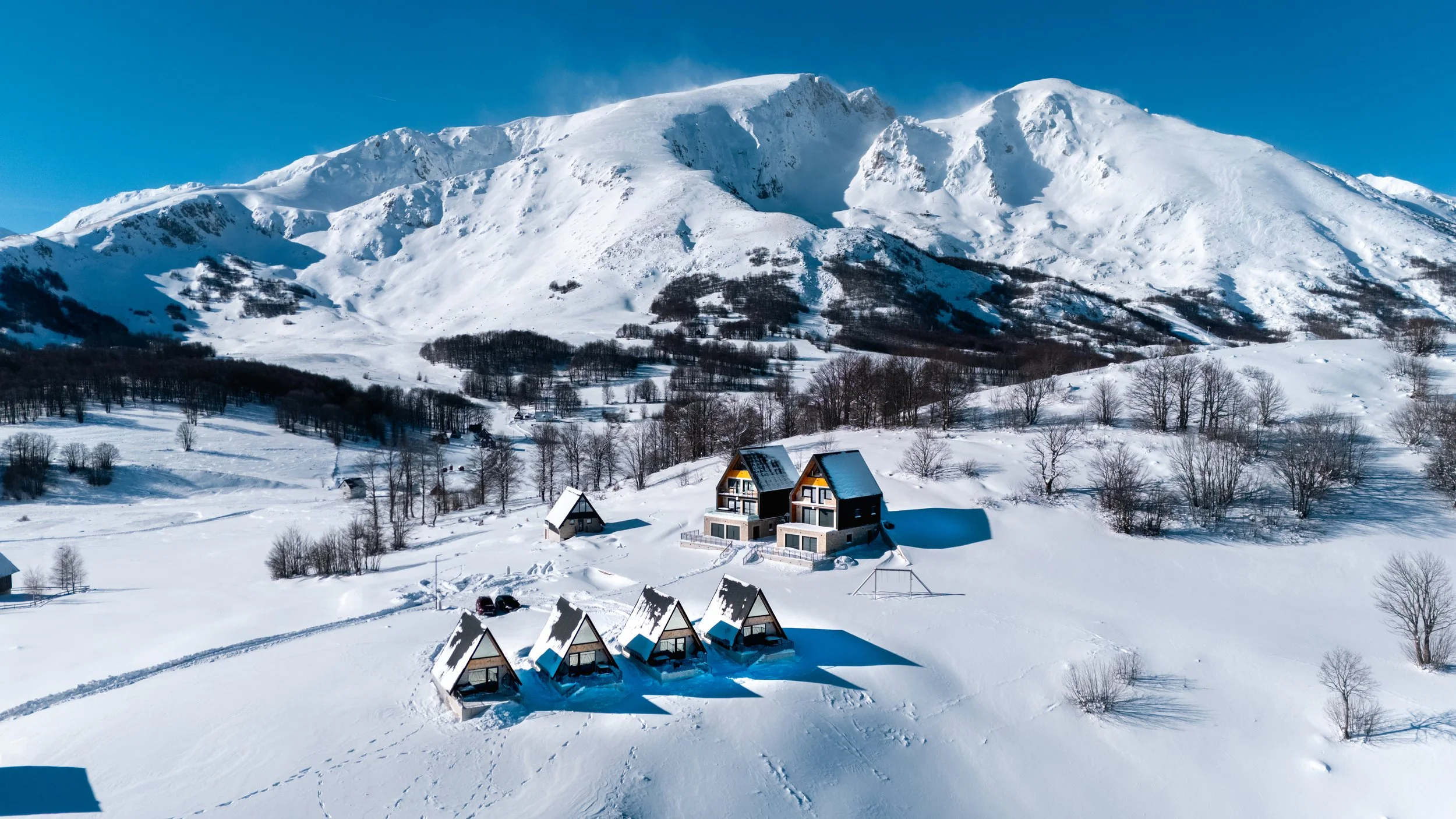 Snow-covered mountains with several houses at the base, surrounded by leafless trees and a clear blue sky.