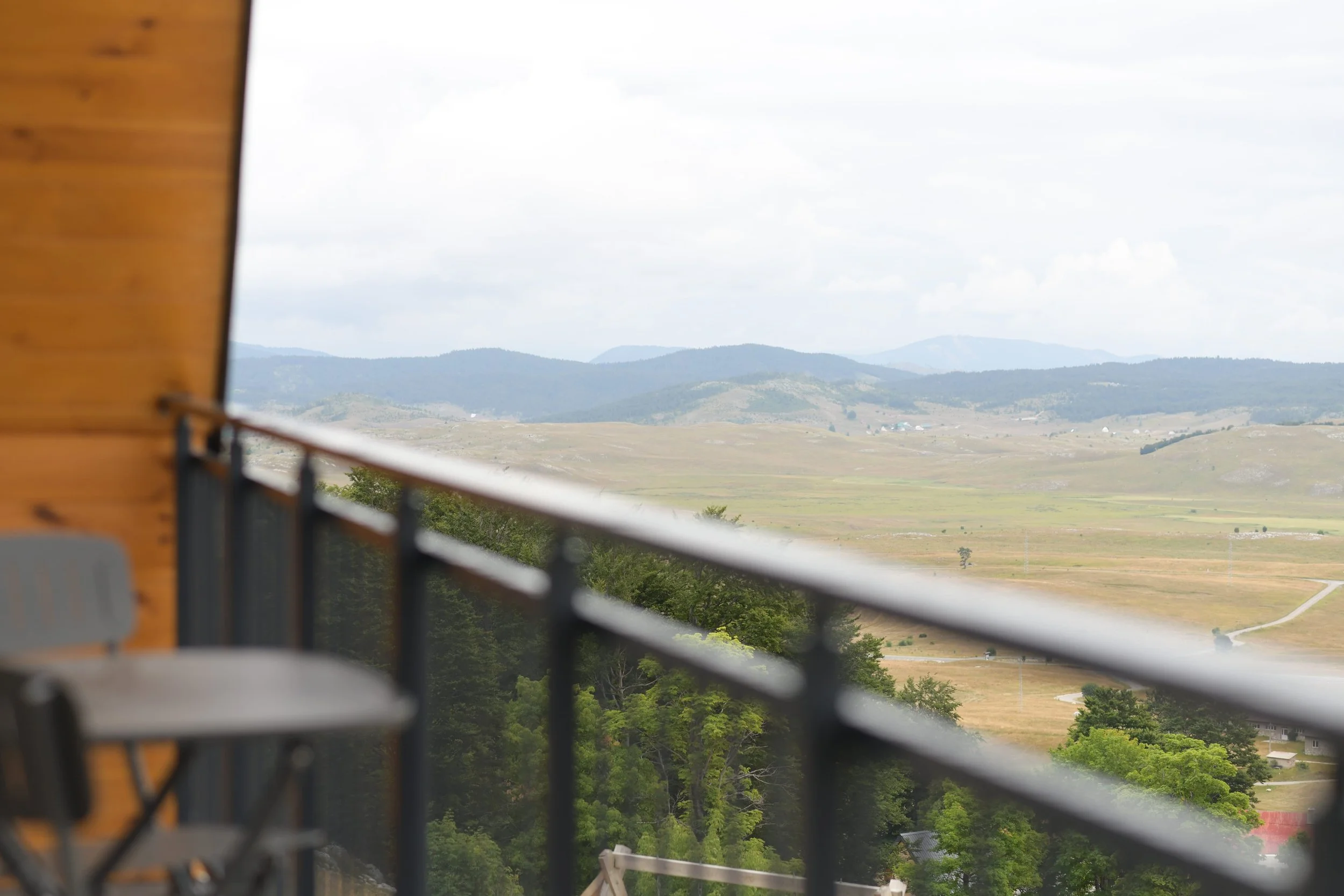 View of rolling hills and open fields seen from a balcony with a black metal railing, part of a wooden structure on the left, under a cloudy sky.