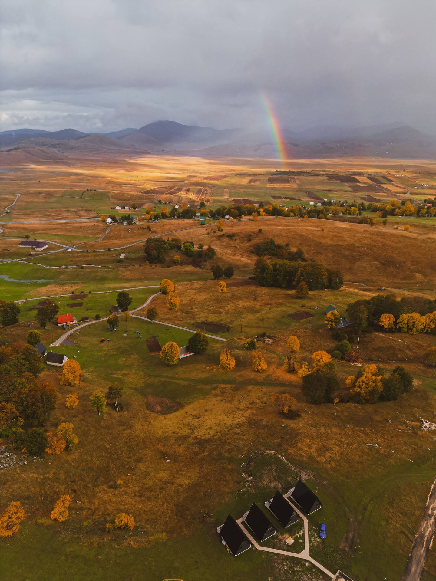 A panoramic aerial view of a rural landscape featuring a rainbow, rolling fields, scattered houses, and mountains in the background during fall.