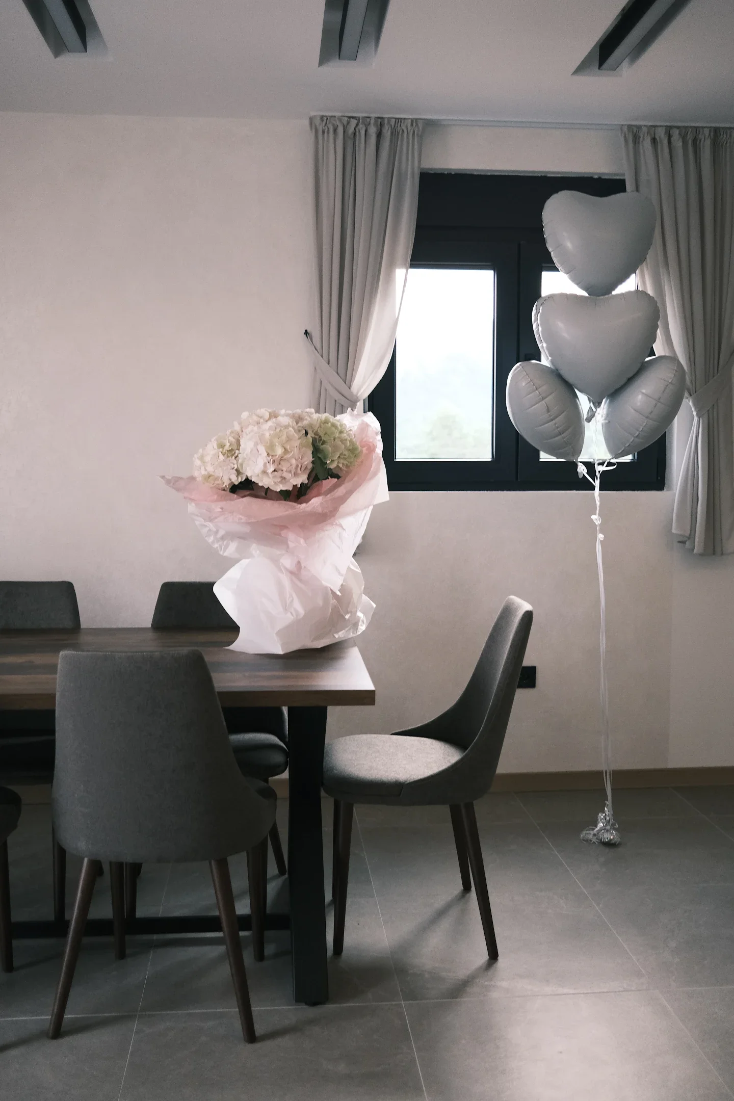 A dining room with a bouquet of white and pink flowers on a wooden table and a bunch of silver heart-shaped balloons next to it, set near a window with cream-colored curtains.