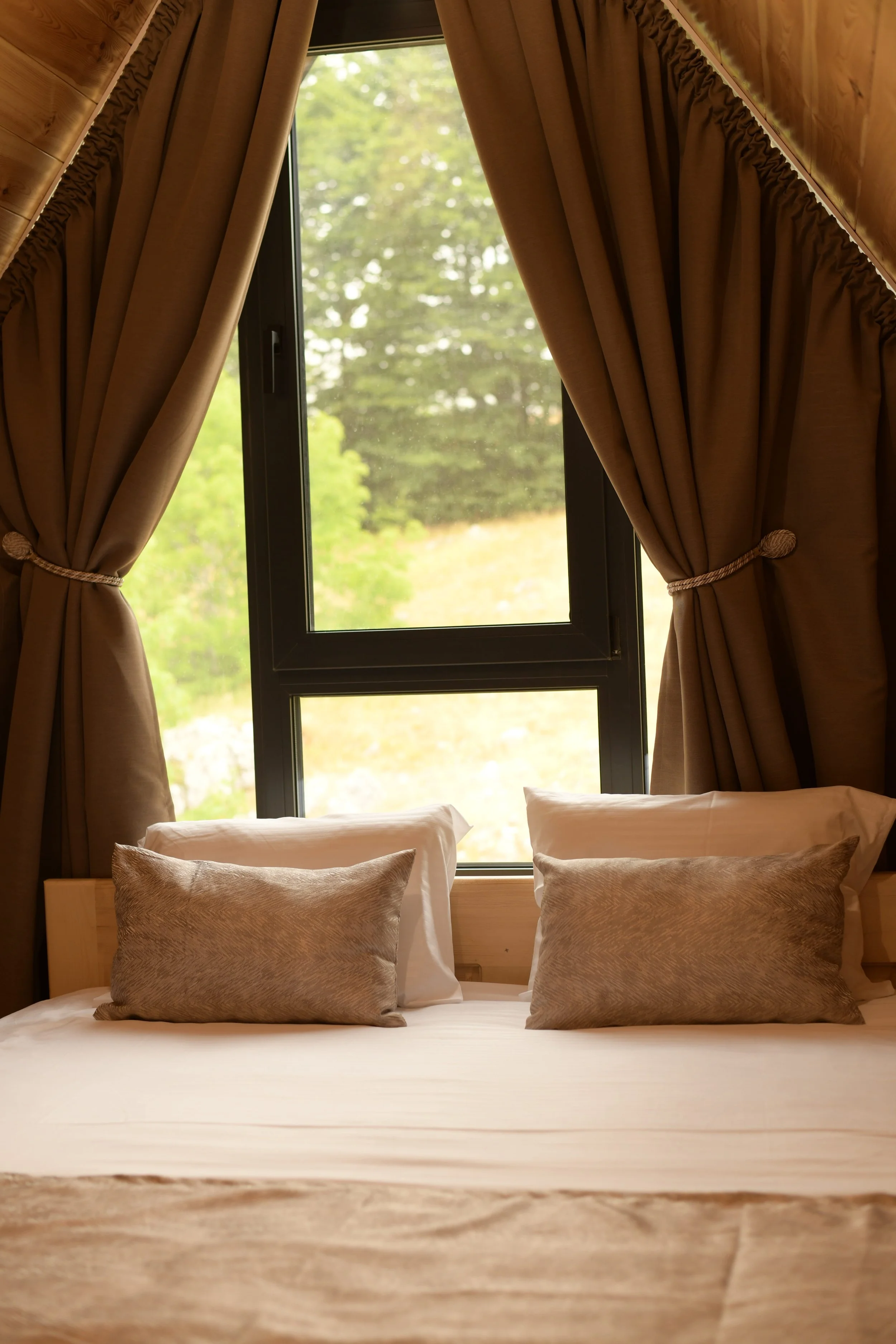 A cozy bedroom with a neatly made bed, gray pillows, and a window with brown curtains revealing a view of green trees outside.