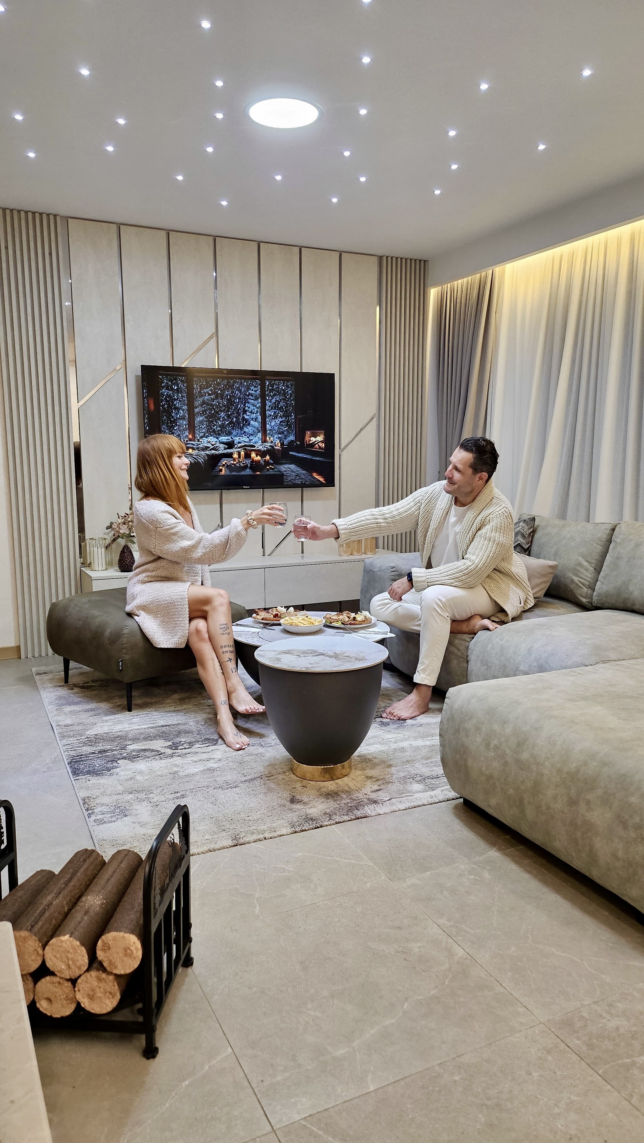 A couple sitting on sofas in a modern living room, clinking glasses and enjoying snacks, with a large TV behind them and a wood-decorated wall, cozy lighting, and a wood log holder on the floor.