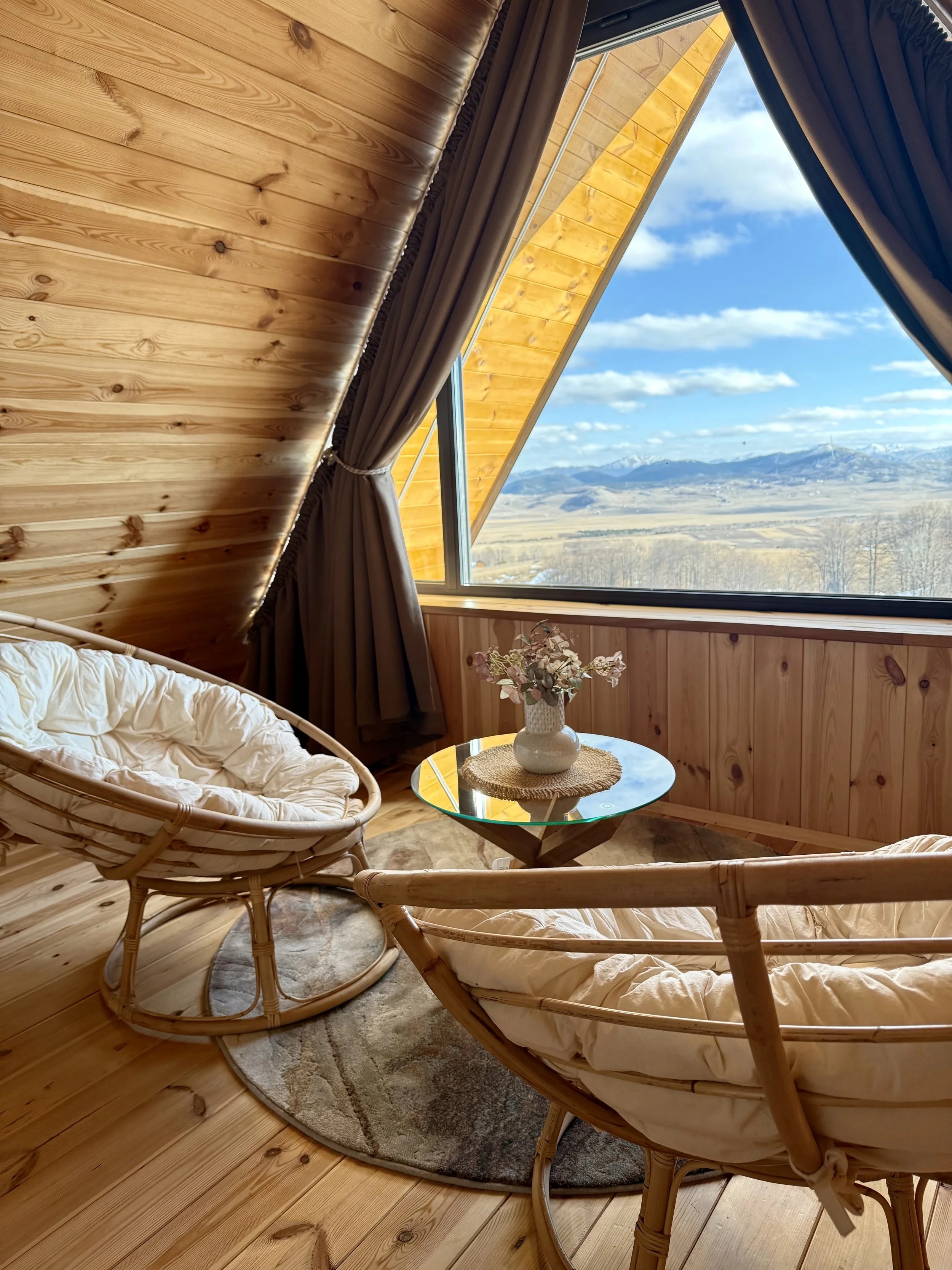 Cozy room with wooden walls and ceiling, featuring a large triangular window with a view of mountains and a blue sky, two rattan chairs, a glass-top table with a floral vase, and a beige rug.