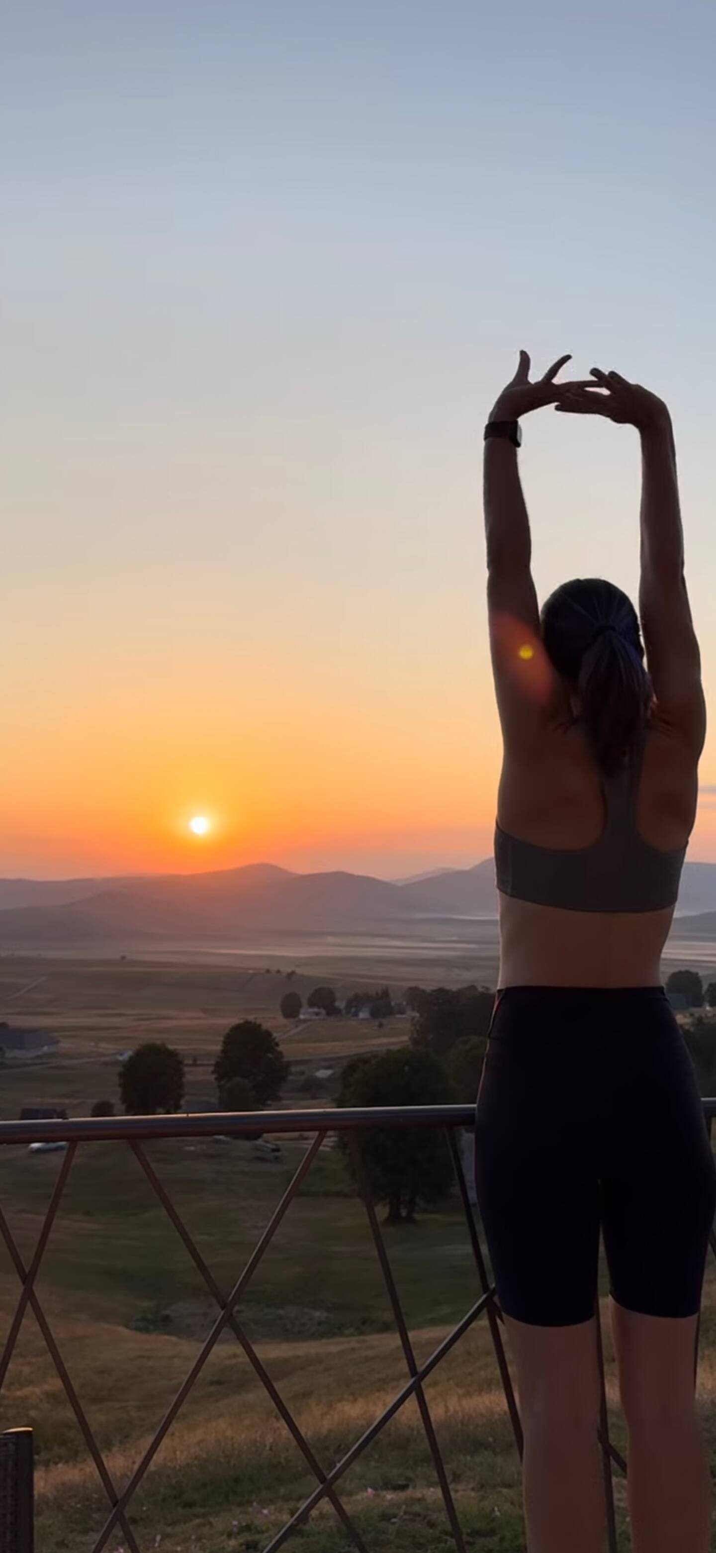 A woman in athletic wear stretching on a balcony during sunset over a hilly landscape with trees.